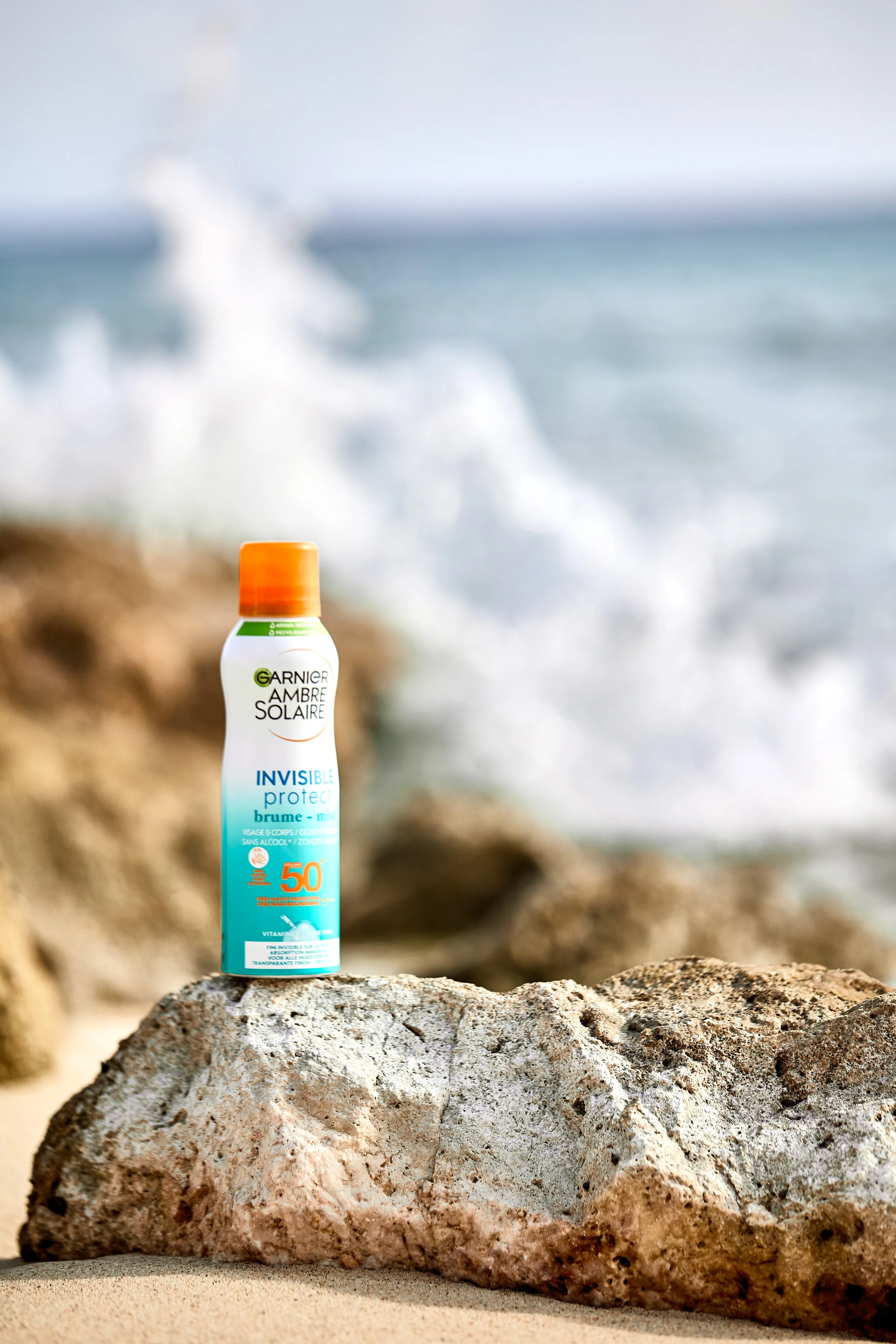 A bottle of Garnier Ambre Solaire Invisible Protect spray sunscreen with SPF 50 on a rock at the beach, with ocean waves in the background.