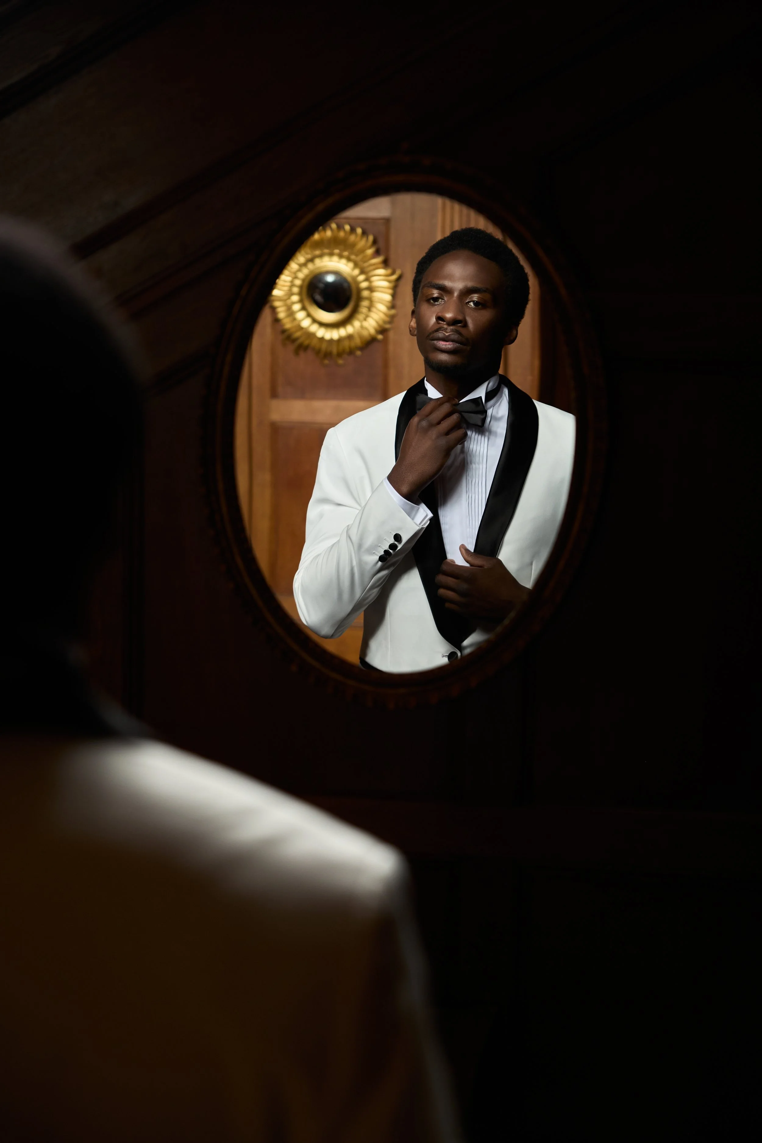 A man in a white tuxedo with a black bow tie looking at himself in a mirror adjusting his bow tie.