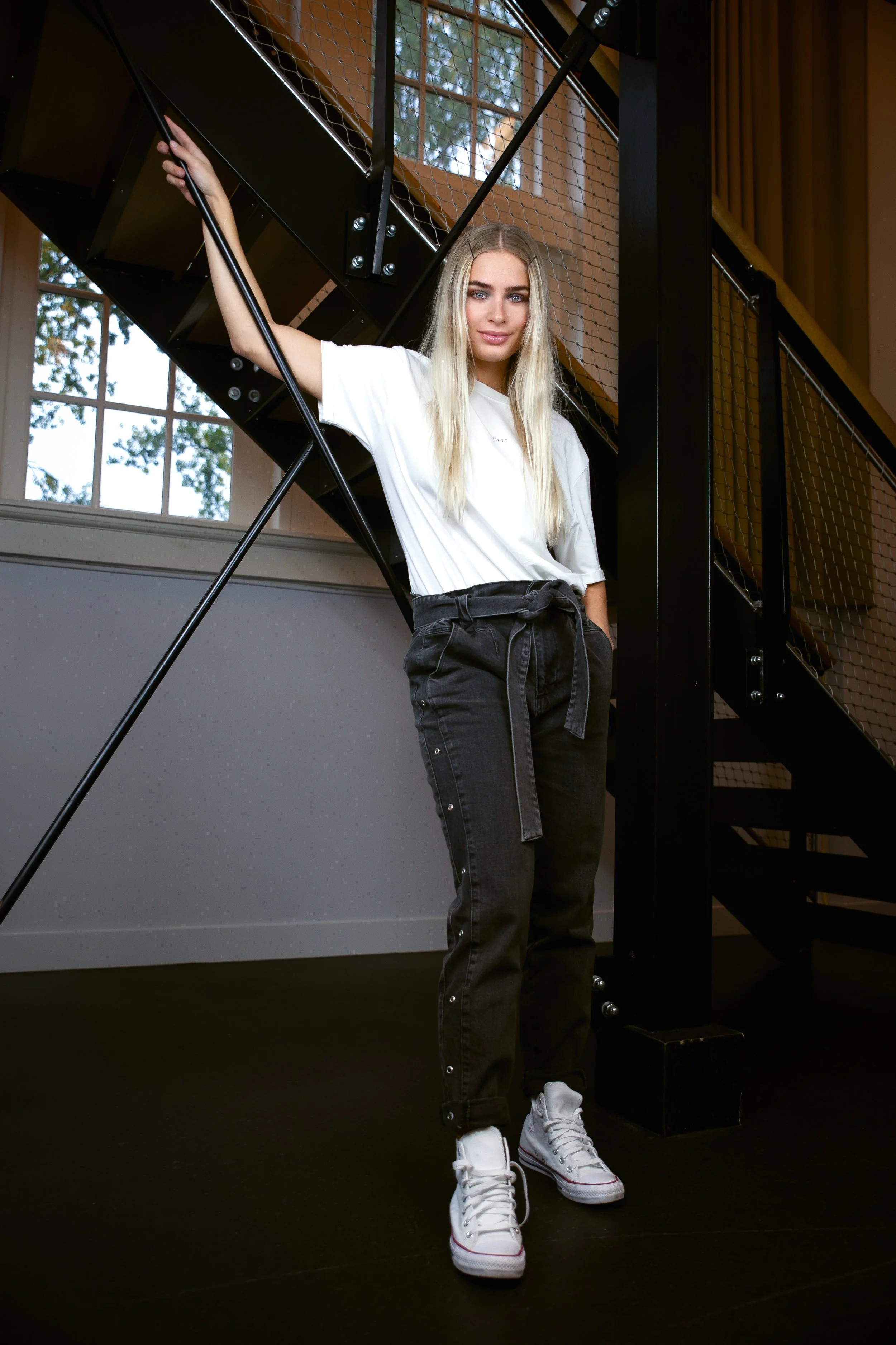 A young woman with long blonde hair and blue eyes stands on a black floor near a staircase, wearing a white T-shirt, black pants with a tied belt, and white sneakers, indoors with large windows behind her.
