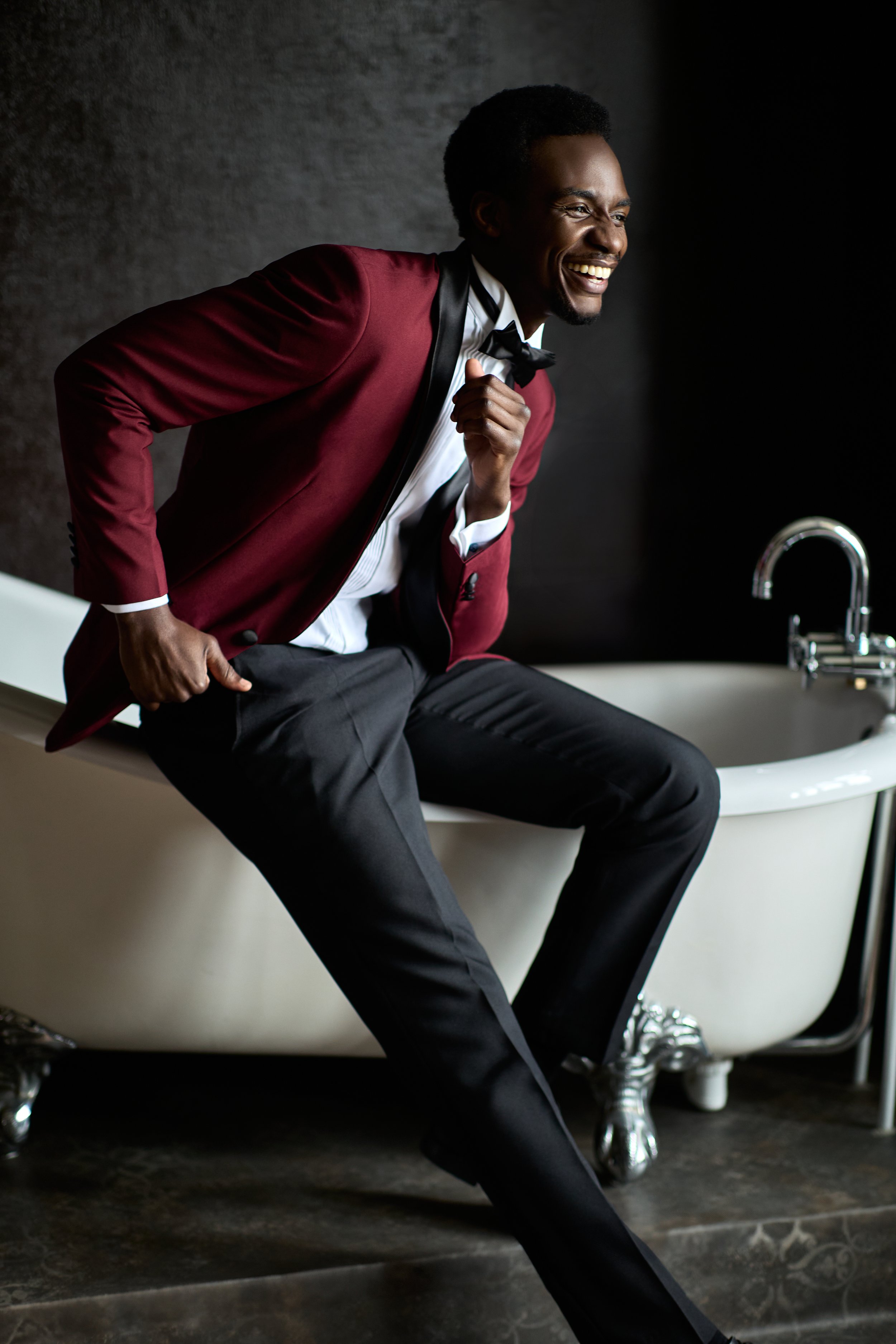 A man in a tuxedo with a red blazer and black bow tie sitting on the edge of a vintage bathtub, smiling and appearing to be laughing.