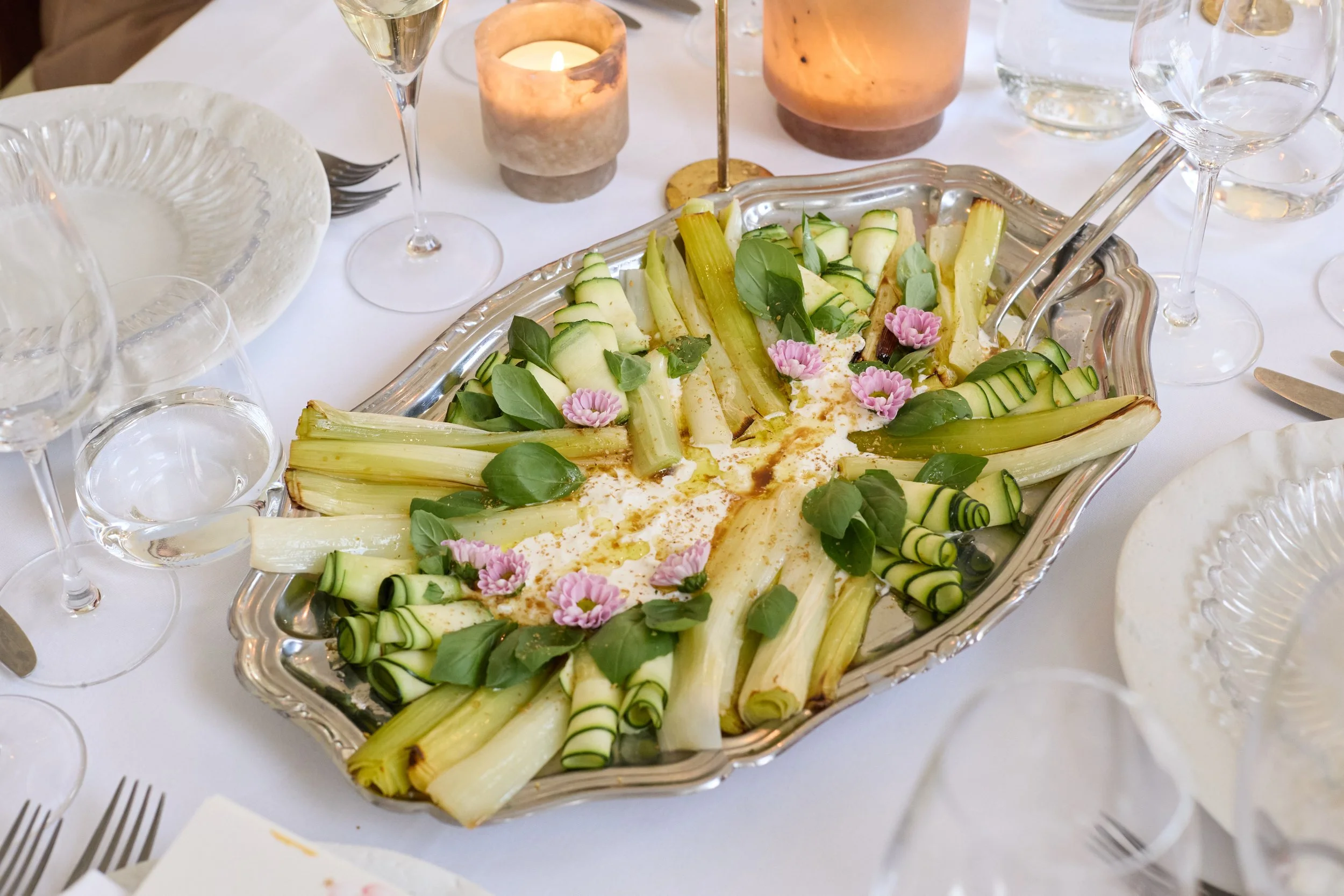 A restaurant table setting with a menu featuring a cute bear on a cloud illustration, a white textured plate, silverware, and a flower arrangement in a glass vase with pink, purple, yellow, and green flowers.