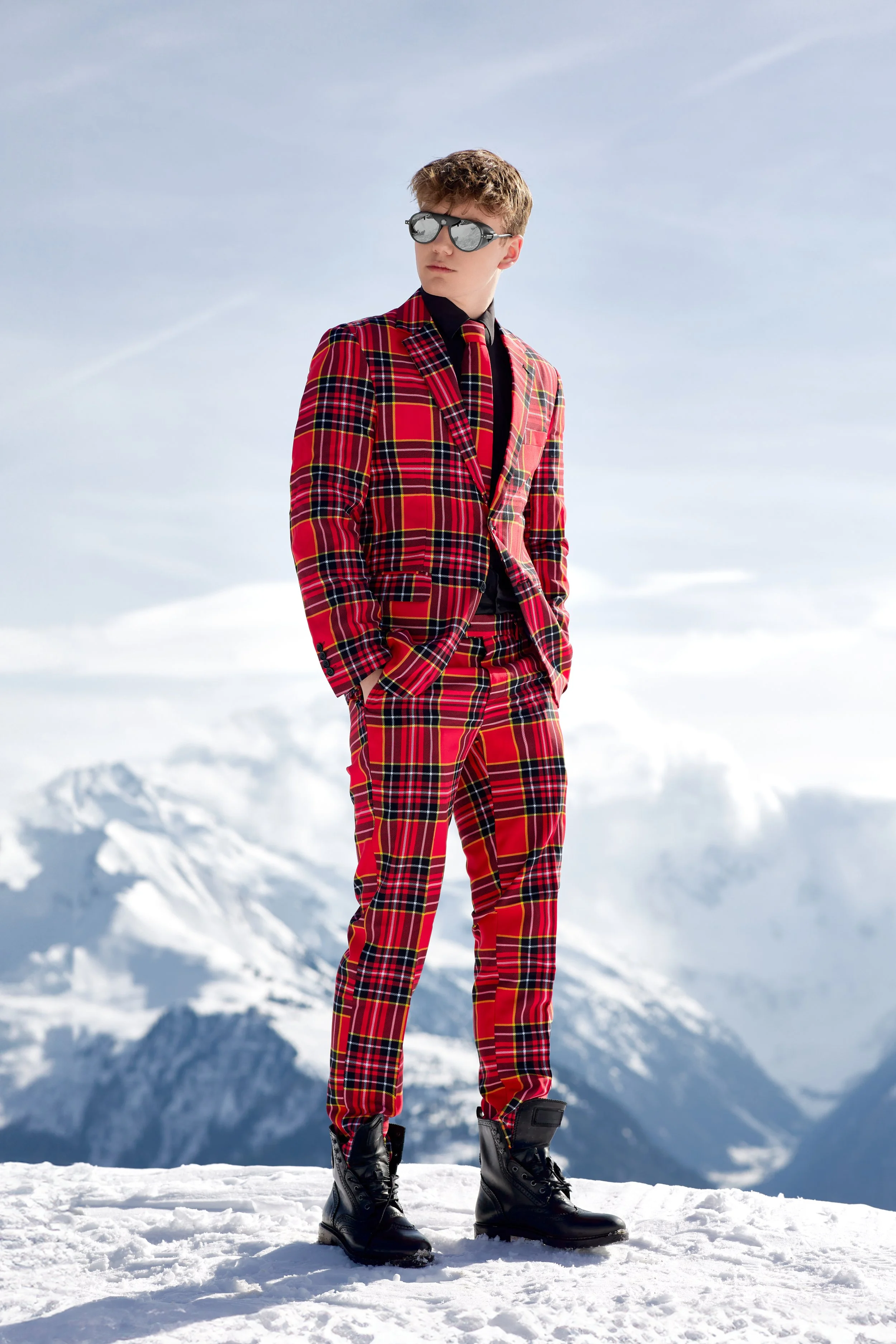 Young man standing outdoors on snow with snow-covered mountains in the background, wearing a red plaid suit, black boots, sunglasses, and a black shirt.