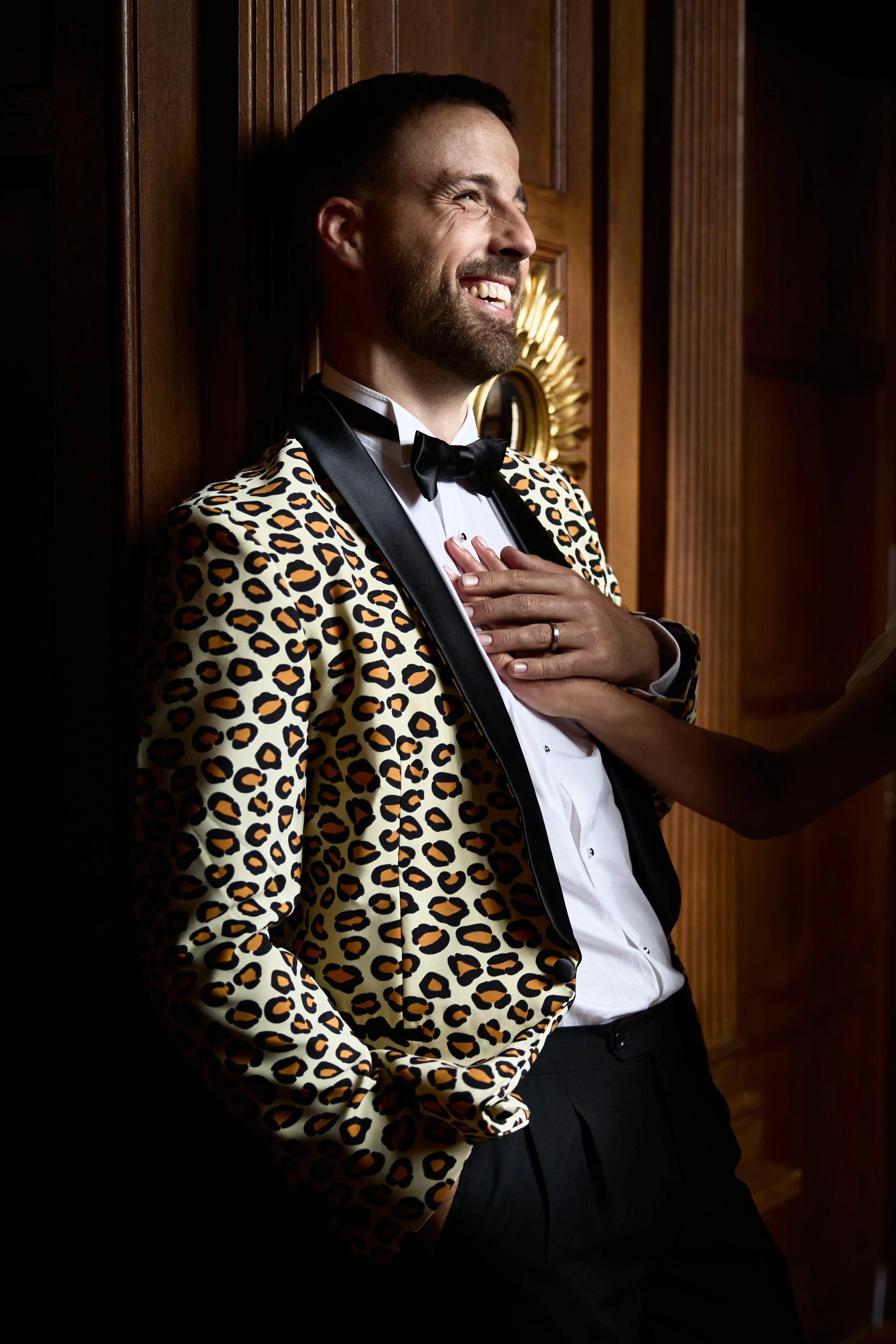 A man in a leopard-print tuxedo jacket and bow tie, smiling and touching his chest, leaning against a wooden wall.