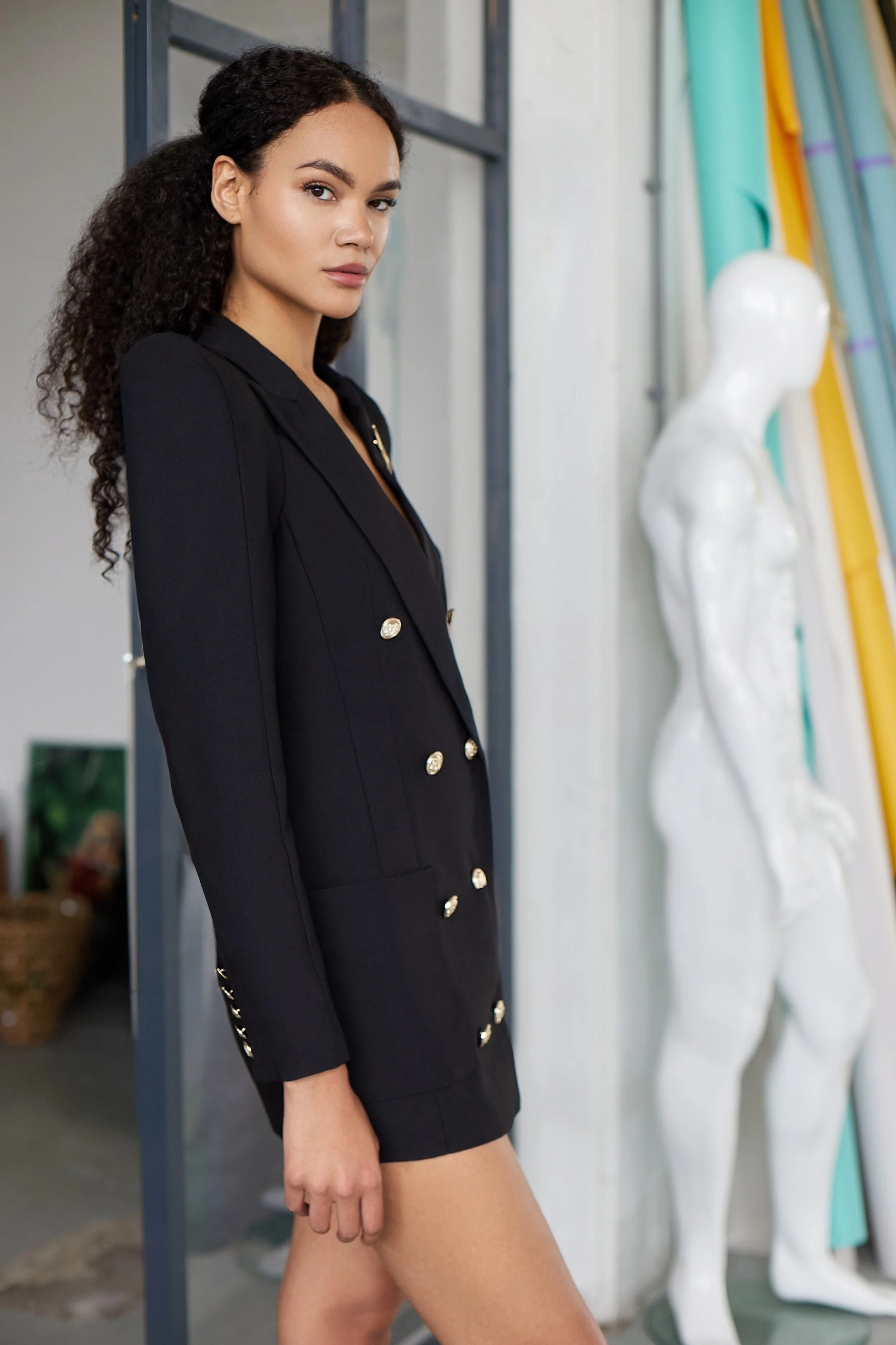 A woman with curly hair wearing a black blazer-dress with gold buttons standing indoors near an art studio mannequin and surfboards.