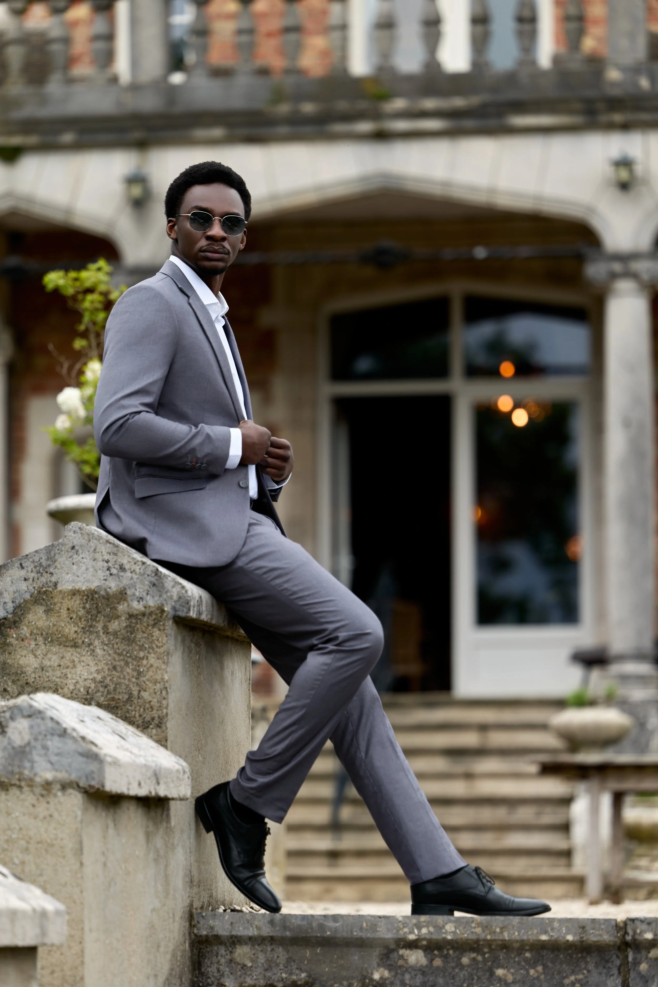 A man in a gray suit with sunglasses sitting on outdoor steps in front of a building with stairs and large windows.