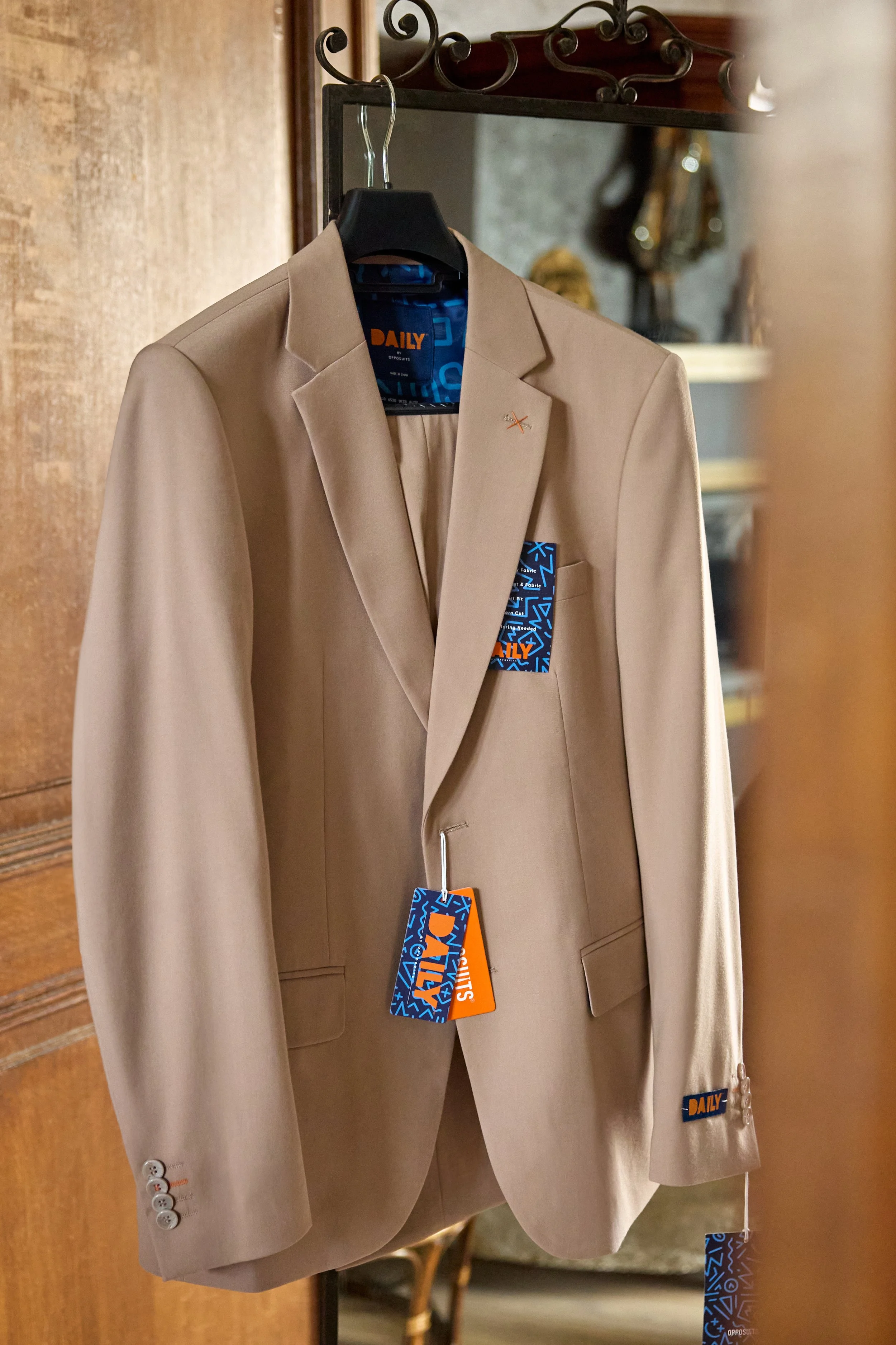 A beige men's suit jacket hanging on a black hanger with tags attached, displayed in front of a mirror.