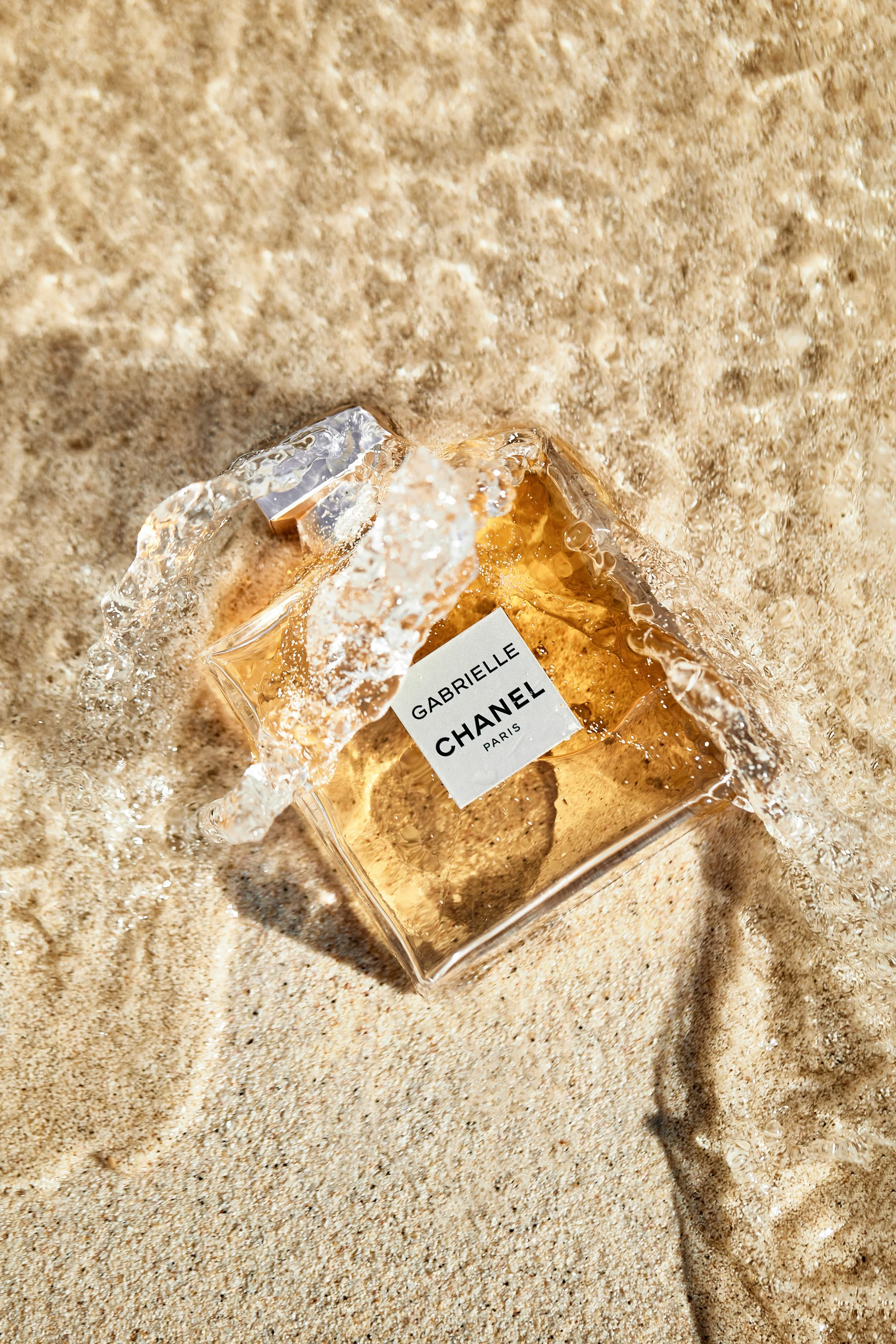 A shattered Chanel perfume bottle labeled Gabrielle on sandy ground with water splashing over it.