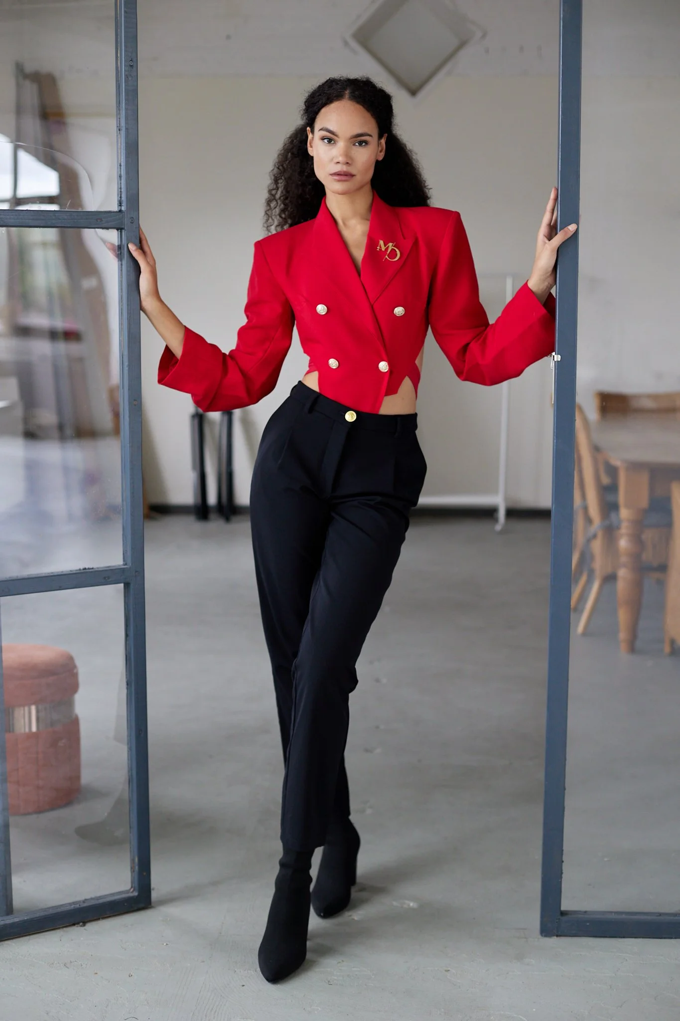 A woman in a red, double-breasted blazer with gold buttons and a gold monogram pins stands in a modern, industrial-style room. She leans against a doorway with one hand, looking directly at the camera.