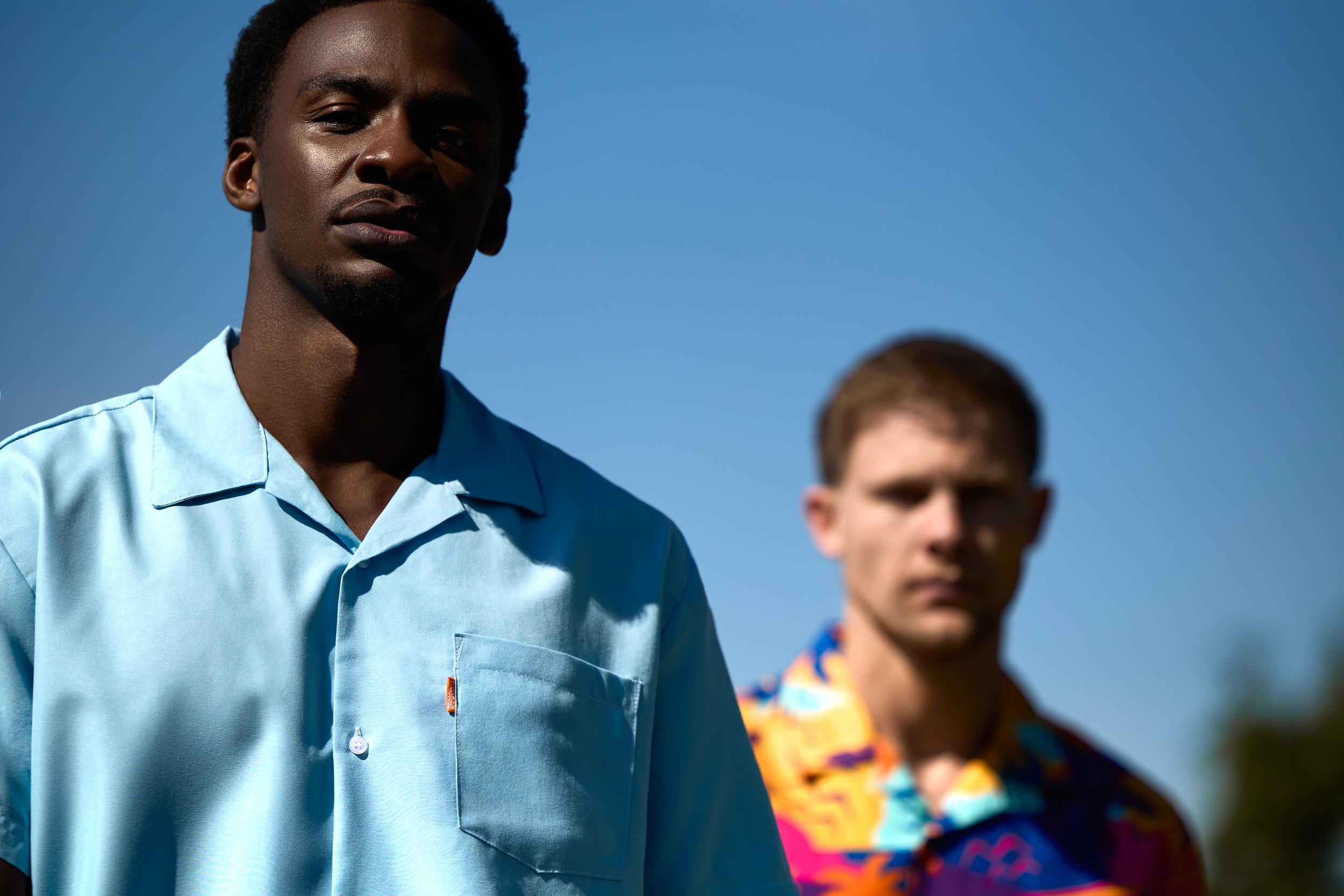 Two men outdoors under a clear blue sky, one in the foreground wearing a light blue shirt, the other in the background wearing a colorful patterned shirt. The man in front has dark skin and short curly hair, while the man behind has light skin and sh