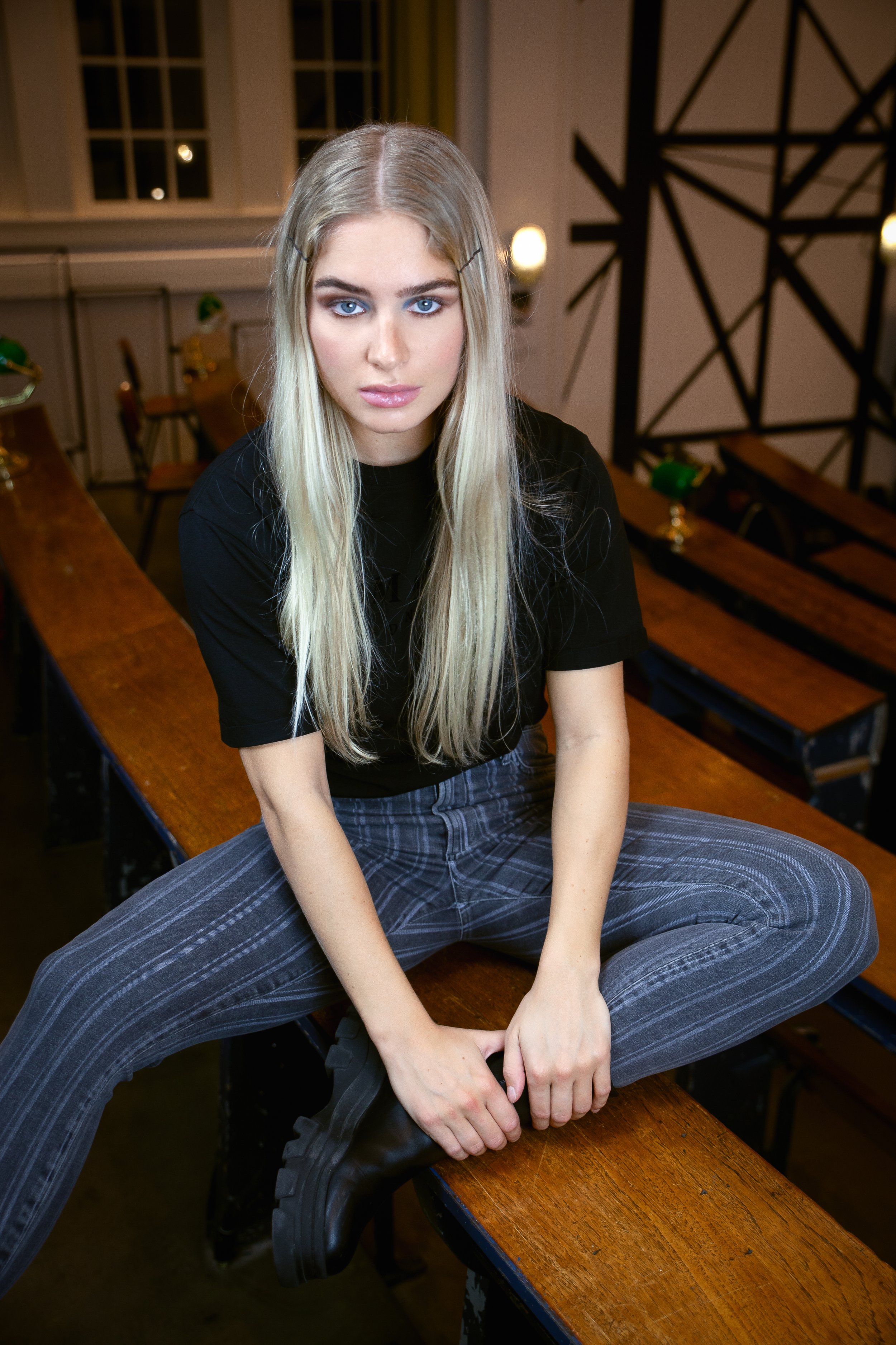 A young woman with long blonde hair and blue eyes sitting on a wooden bench, looking directly at the camera with a serious expression, wearing a black t-shirt, gray striped pants, and black boots.