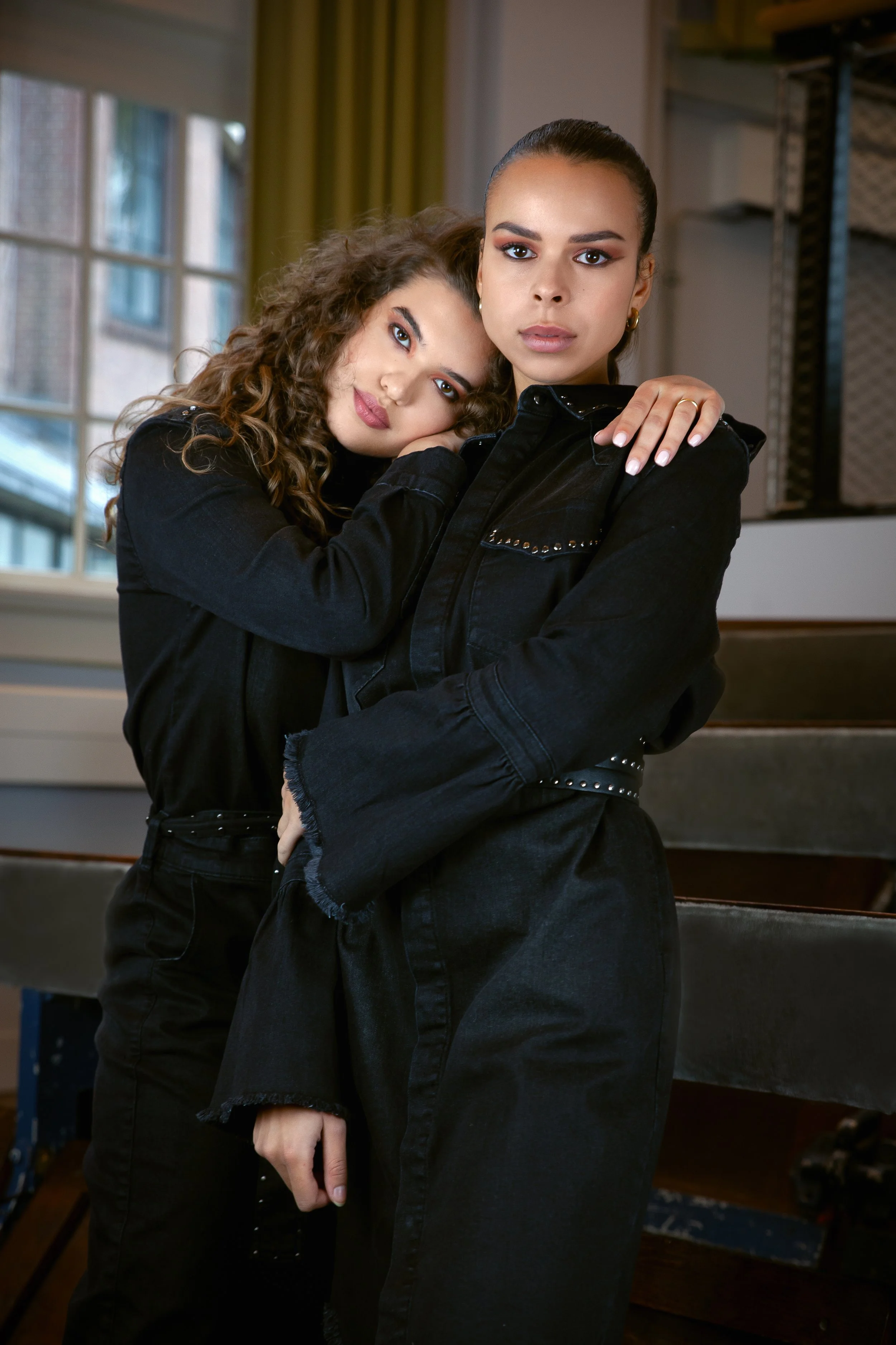 Two women wearing black denim outfits, embracing each other indoors near a window.