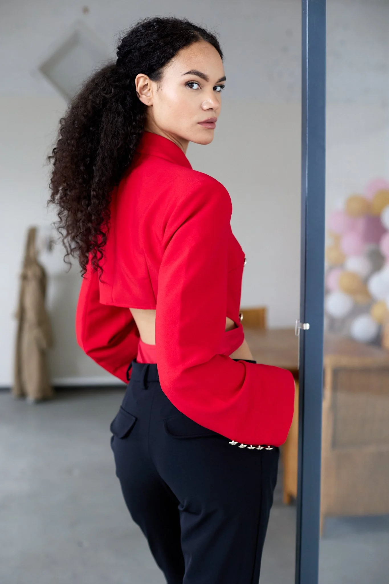 A woman with curly dark hair wearing a red blazer and black pants standing behind a glass display case.