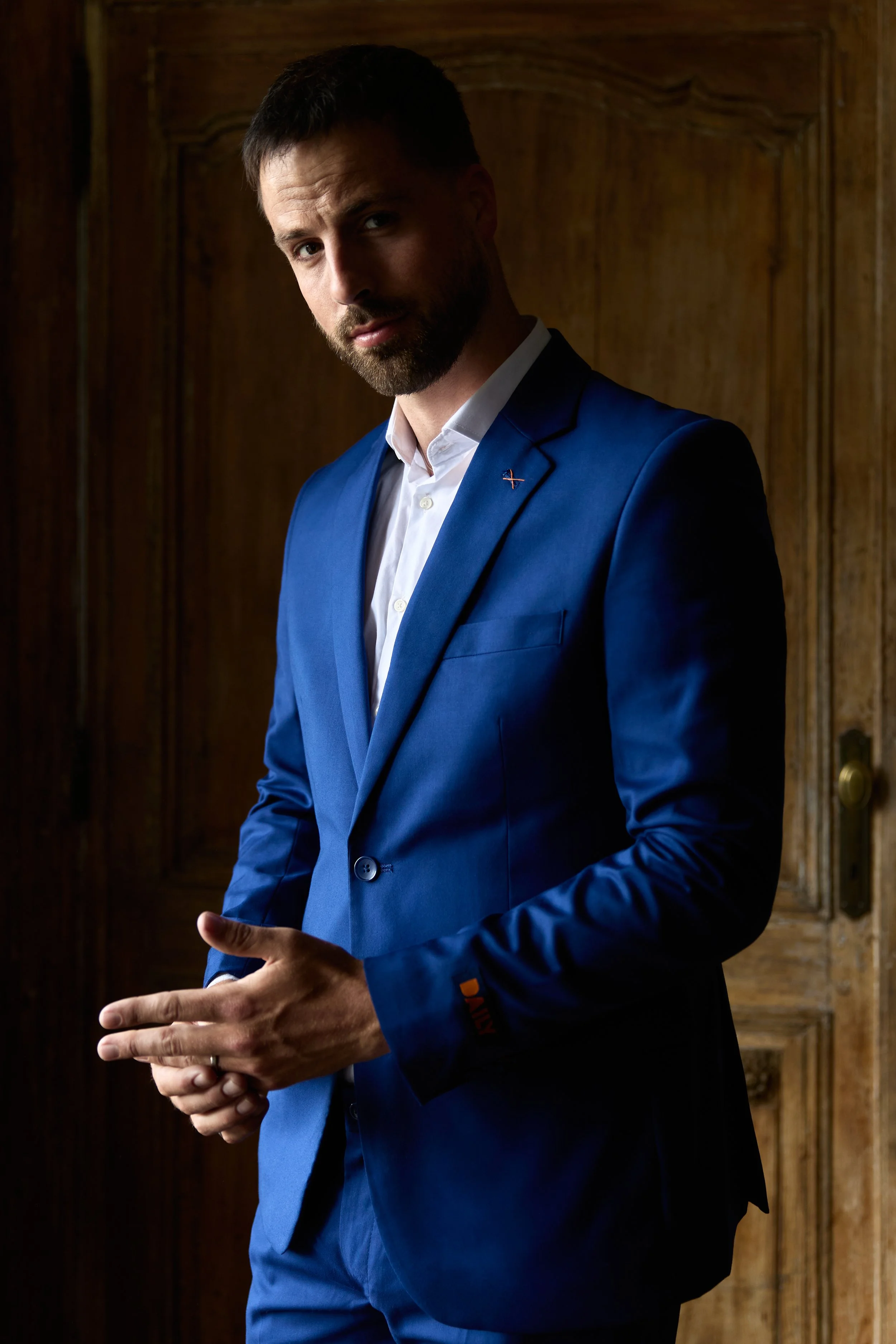 A man in a blue suit and white shirt standing indoors in front of wooden paneling, with a serious expression and his hands clasped.