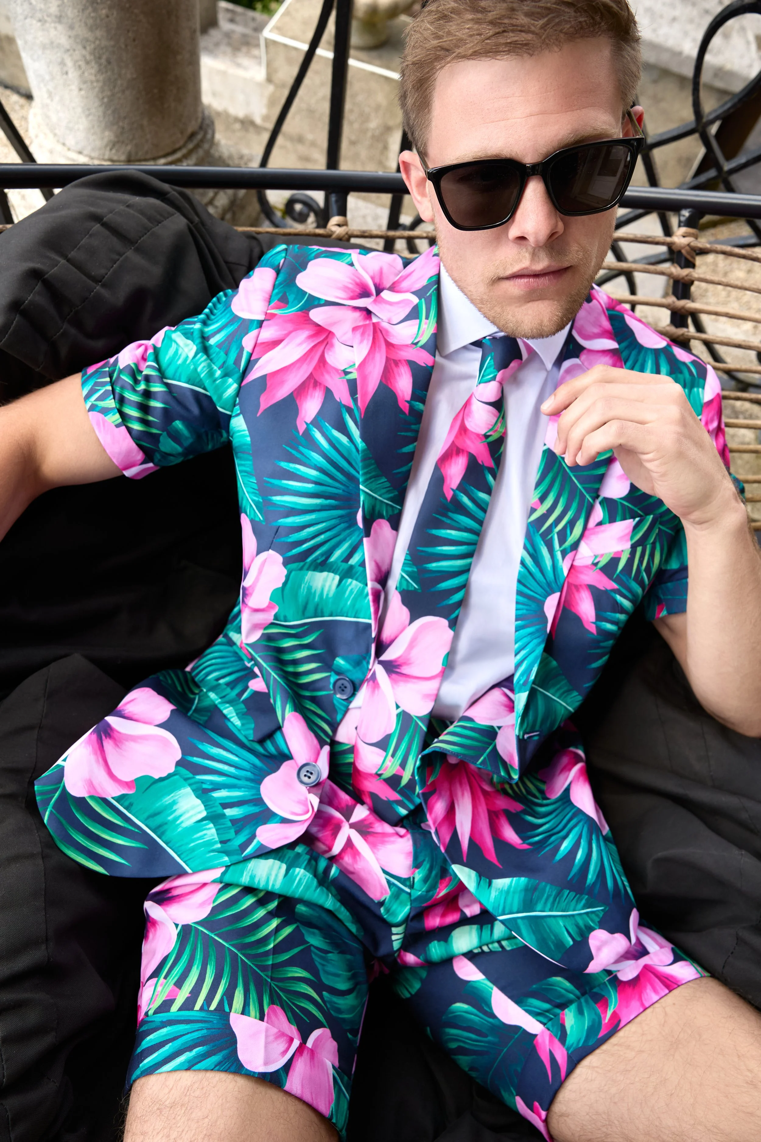 A man wearing a colorful Hawaiian shirt and matching tie, sunglasses, and black pants, sitting outdoors on a bench, with a black bag resting beside him.