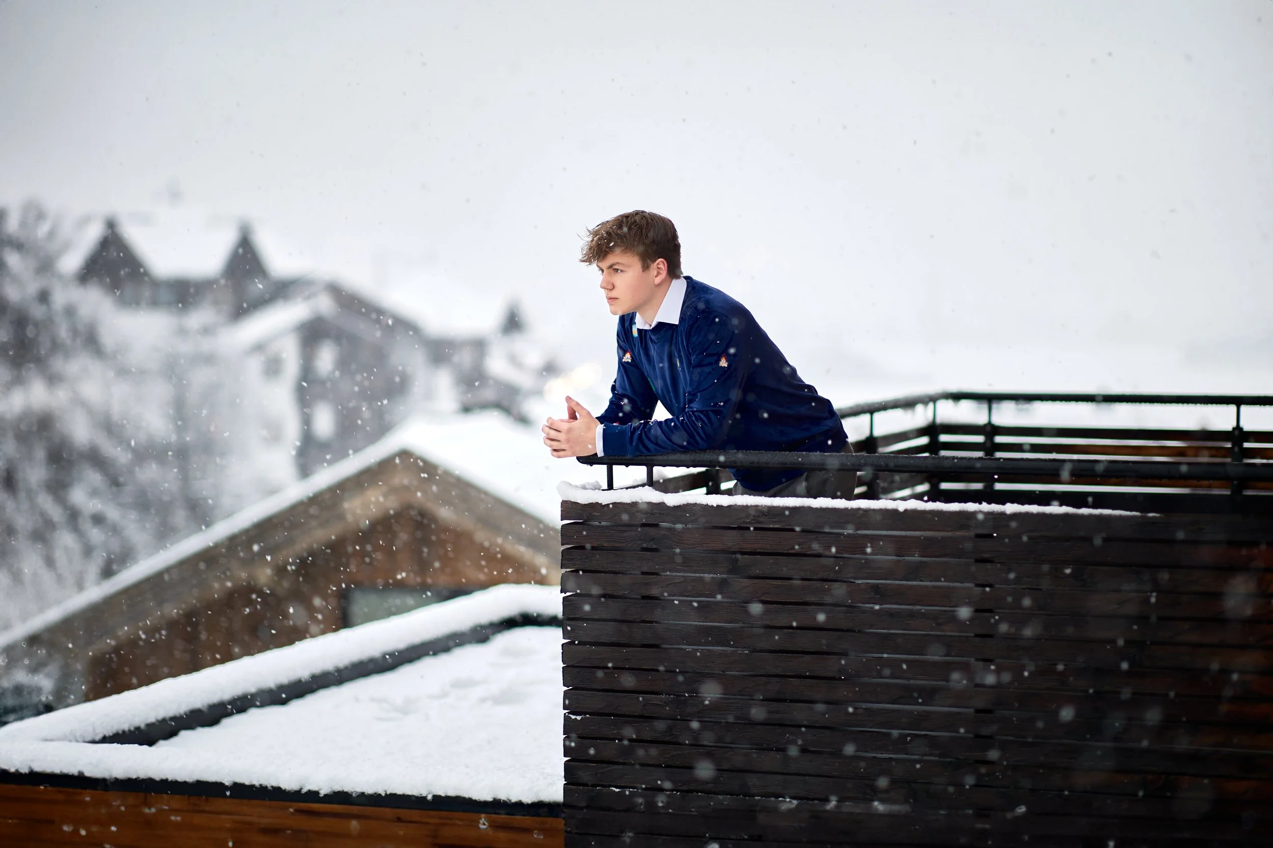 A young man in a blue jacket leaning on a balcony railing outside on a snowy day, looking pensively into the distance with snow falling around him.