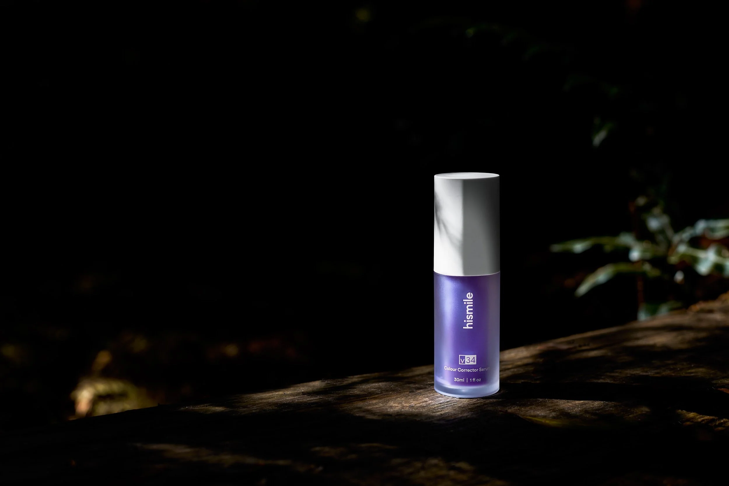 A purple bottle of hismile blue color corrector serum stands on a wooden surface with sunlight casting shadows on it, set against a dark background with faint foliage.