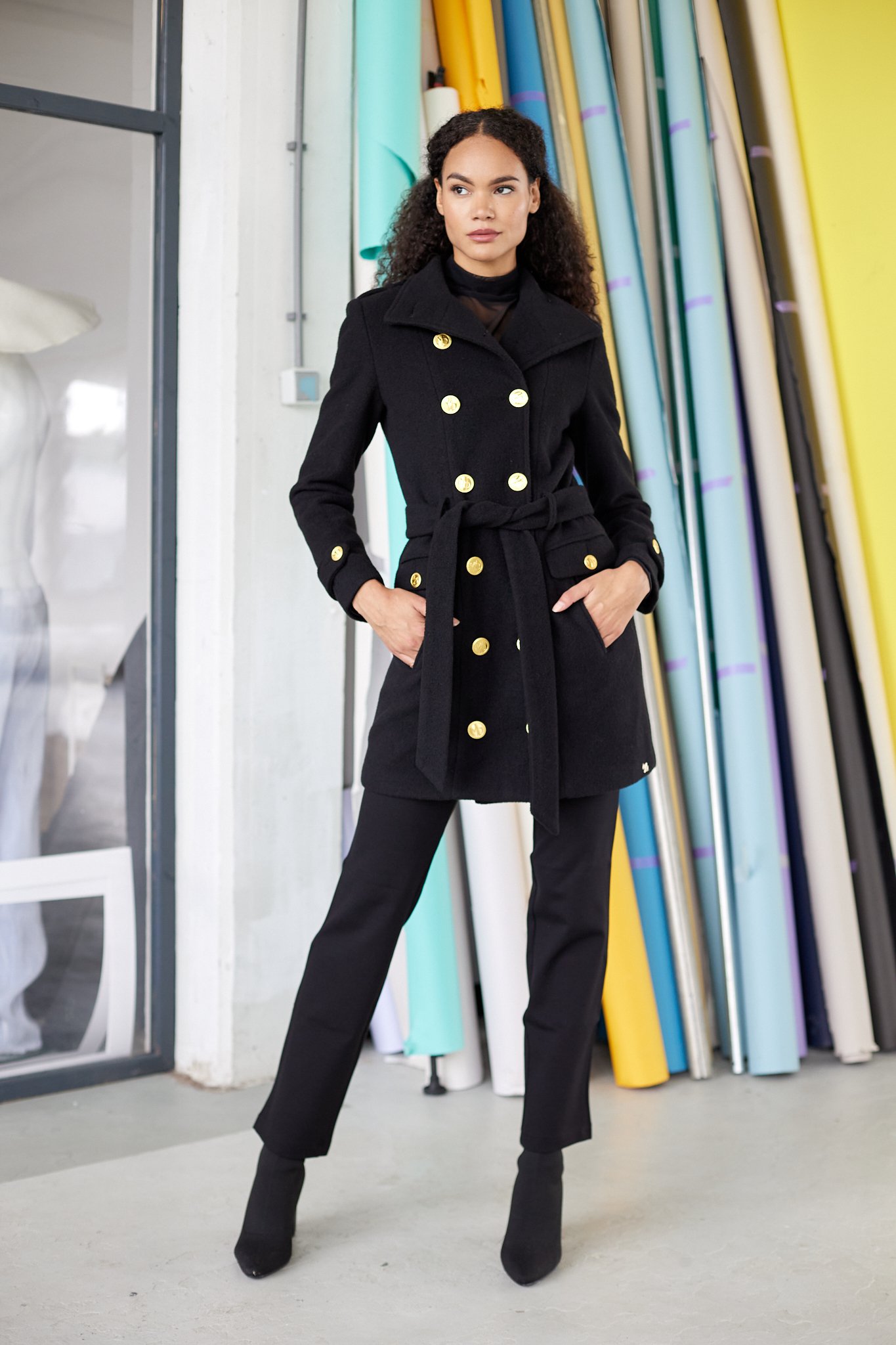 A woman modeling a black coat with gold buttons and a belt, standing indoors with colorful fabric rolls in the background.