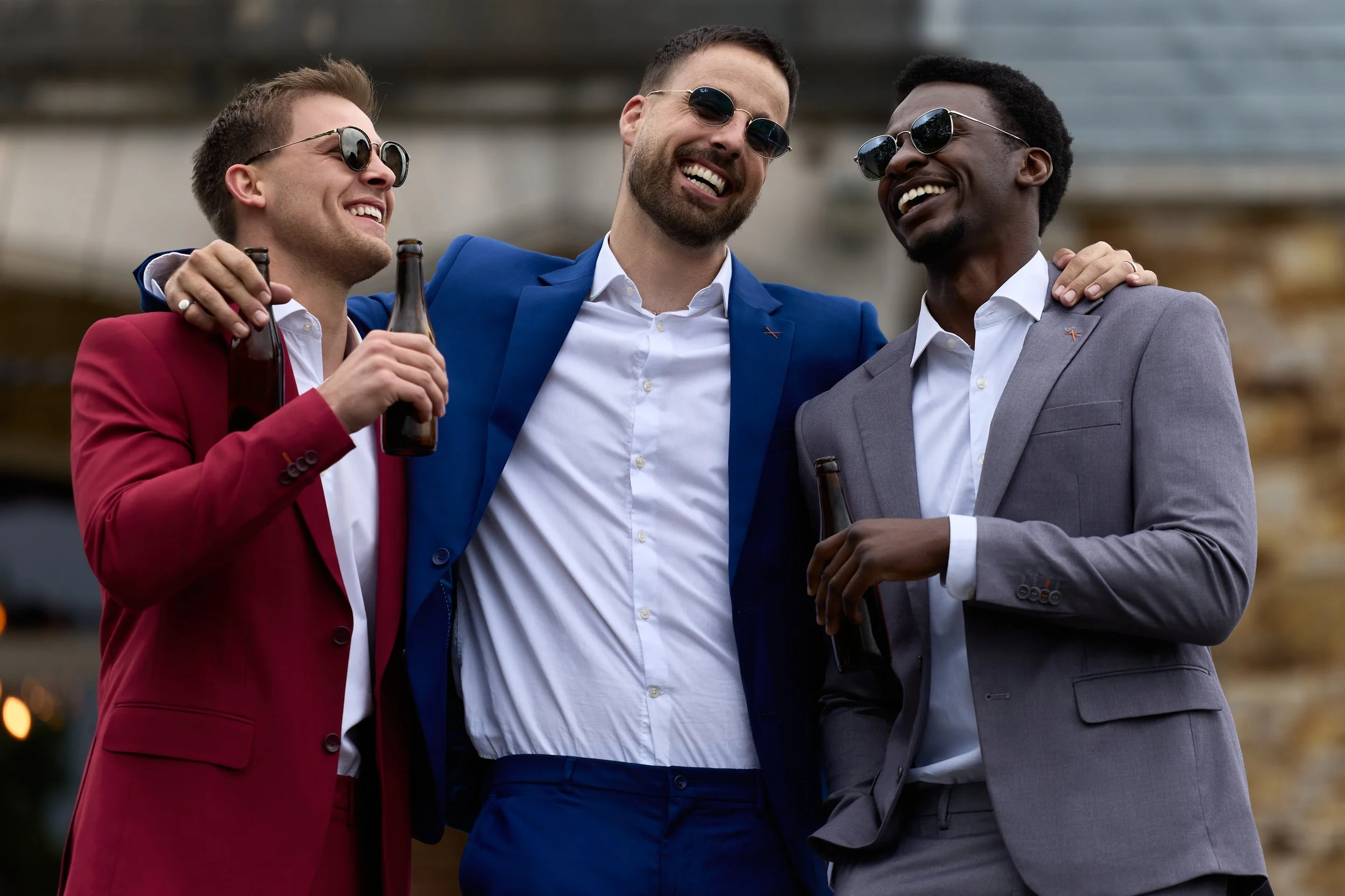 Three men dressed in colorful suits, wearing sunglasses, smiling, and holding beer bottles, standing close together with their arms around each other's shoulders.
