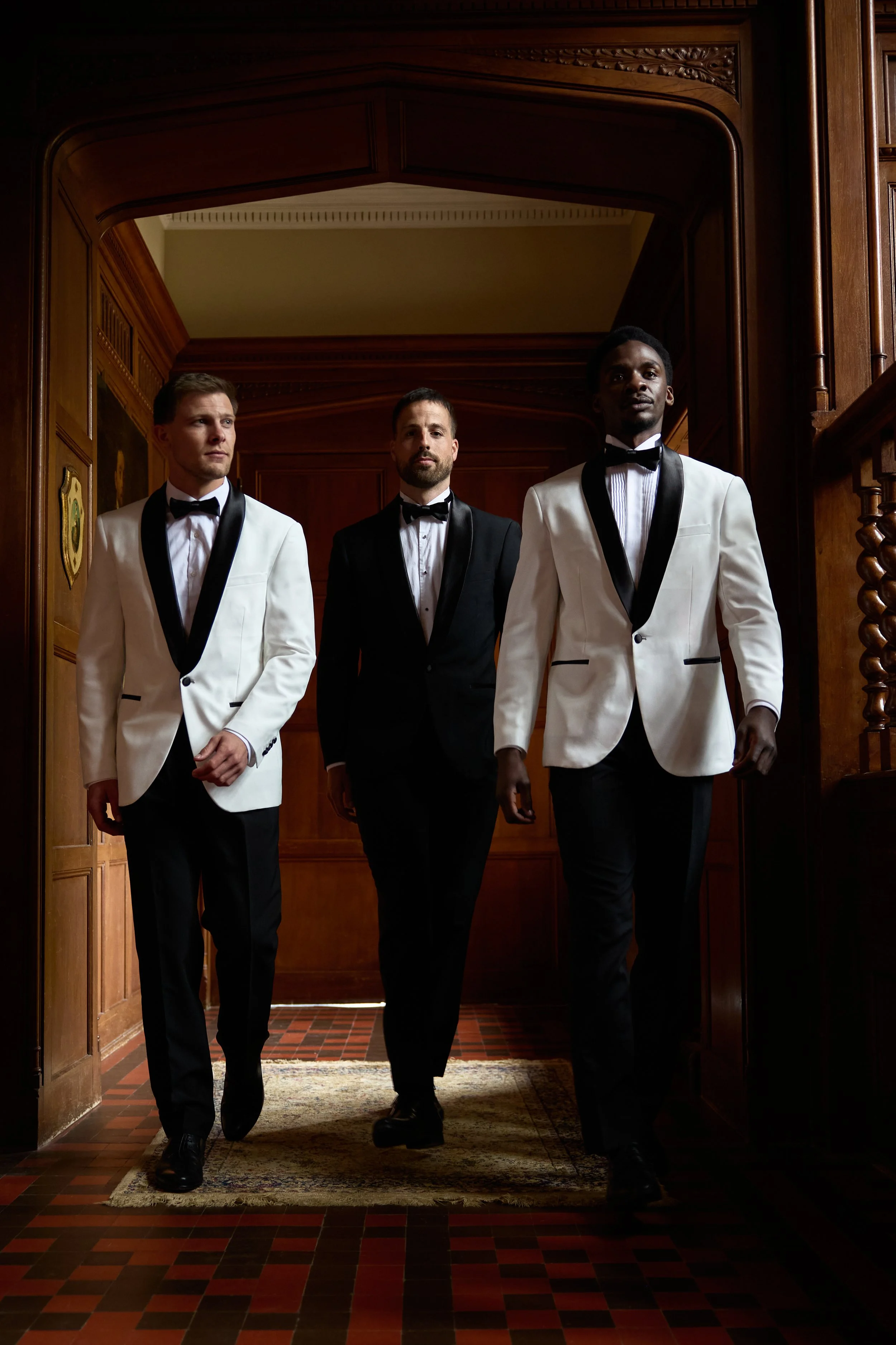Three men walking confidently in a formal setting, dressed in tuxedos with black lapels, white shirts, and black bow ties, inside a wood-paneled hallway.