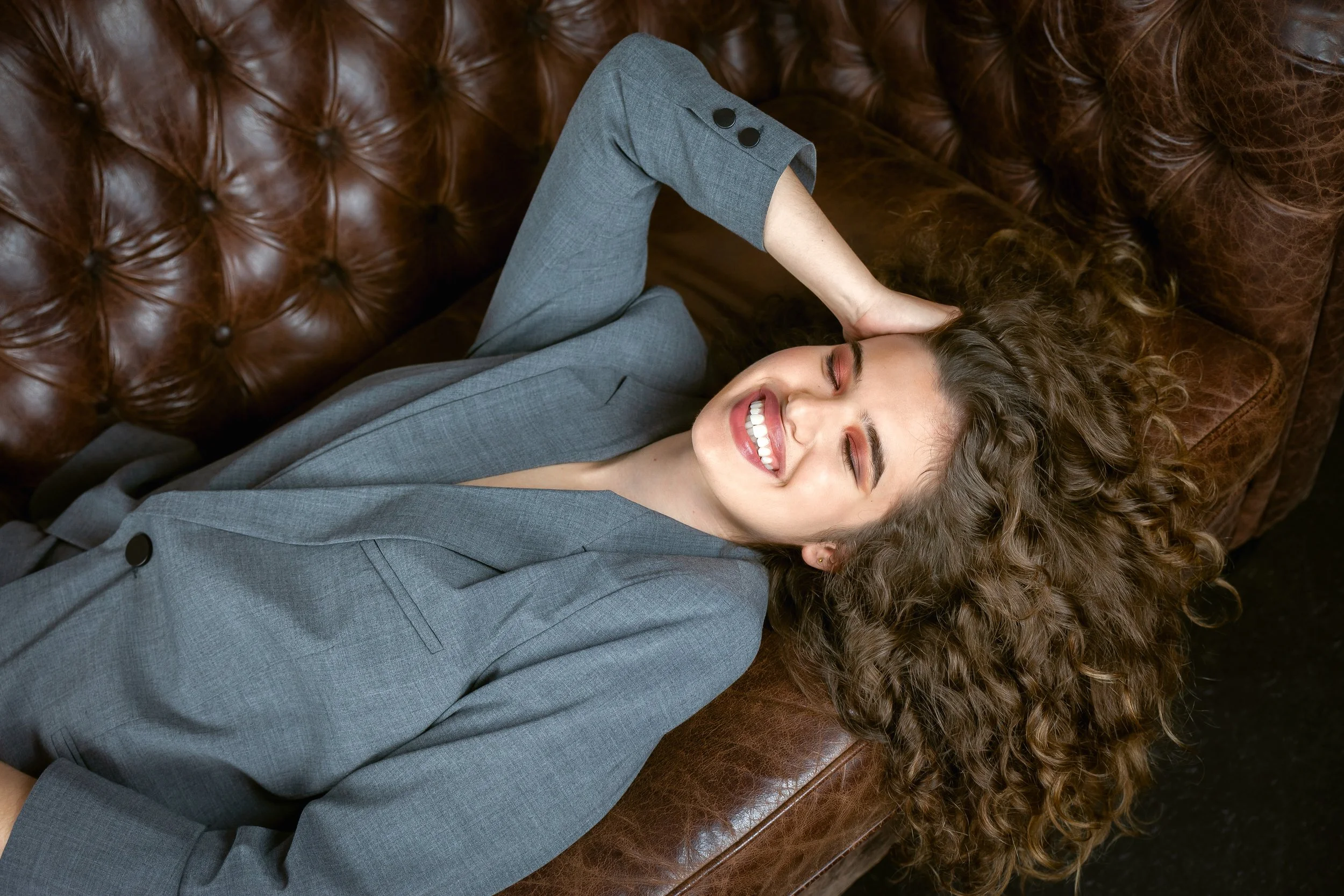 Young woman with curly hair lying on a leather couch, smiling with eyes closed, wearing a gray blazer.