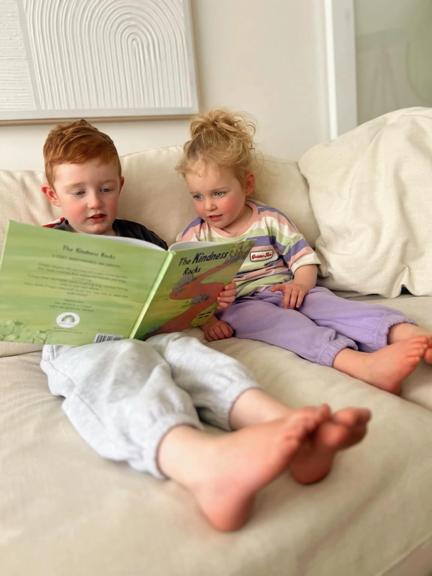 Two young children, a boy with red hair and a girl with curly blonde hair, are sitting on a beige sofa, looking at a green book titled 'The Kindness Rocks'. They are barefoot, and the girl is wearing purple pants. The background includes a white wall with a textured art piece hanging on it.