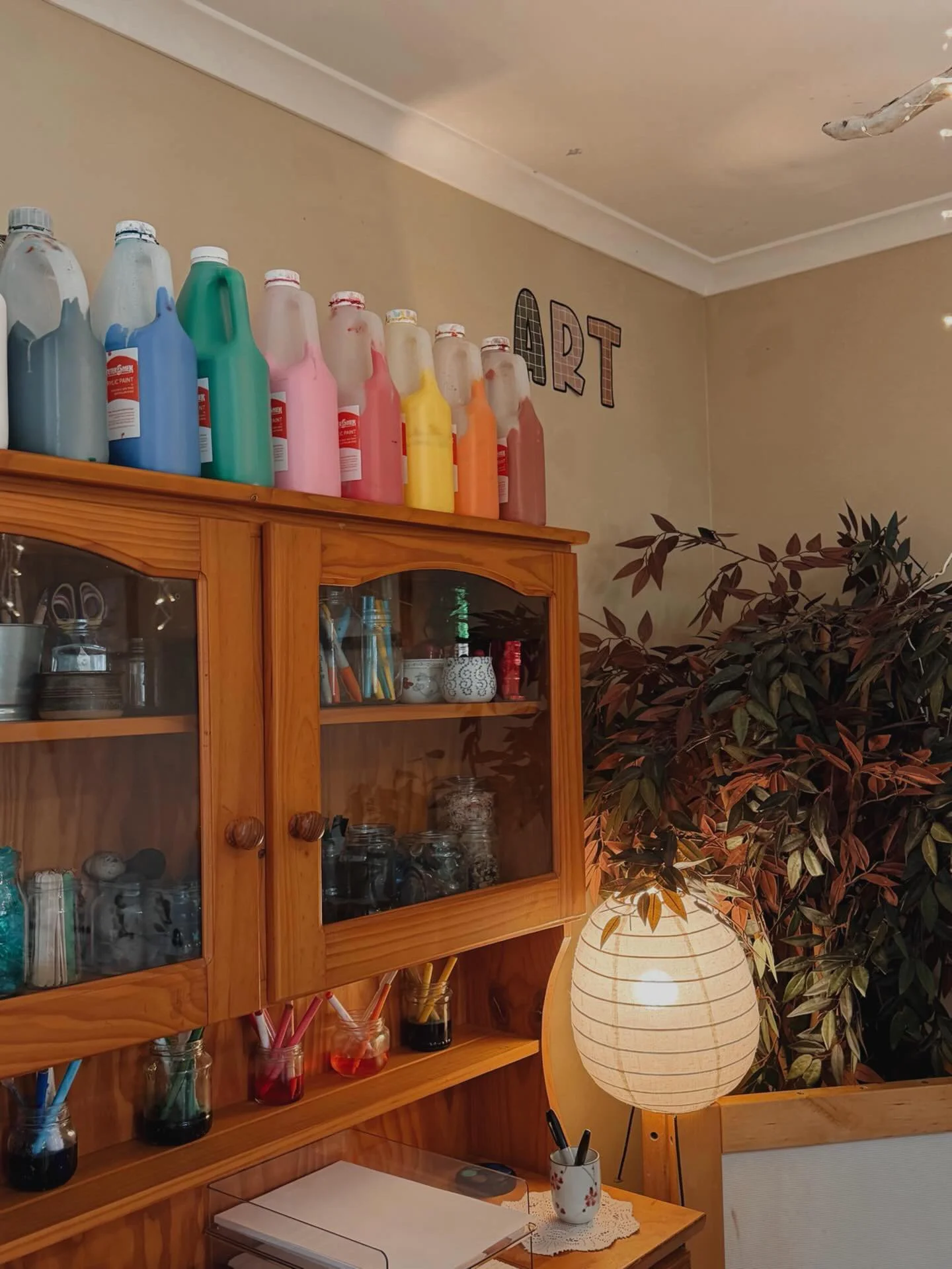 A wooden cabinet with glass doors holding various jars and cups, colorful paint bottles on top, a paper lantern, and a leafy plant in the background.