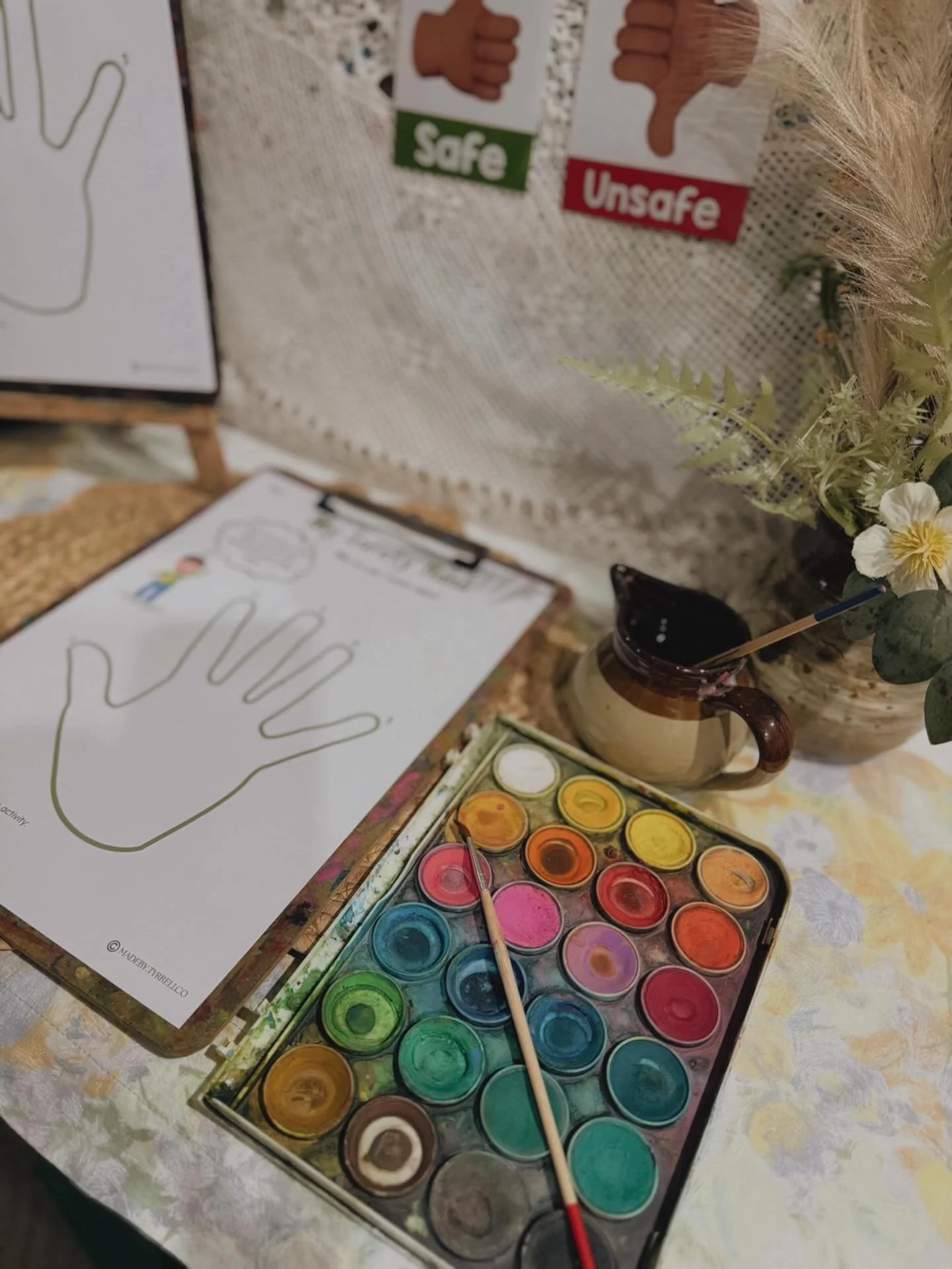 Watercolor paint set with multiple colors, a paintbrush, a partially painted paper with hand outline and safety instructions, a ceramic mug, and flowers on a table.