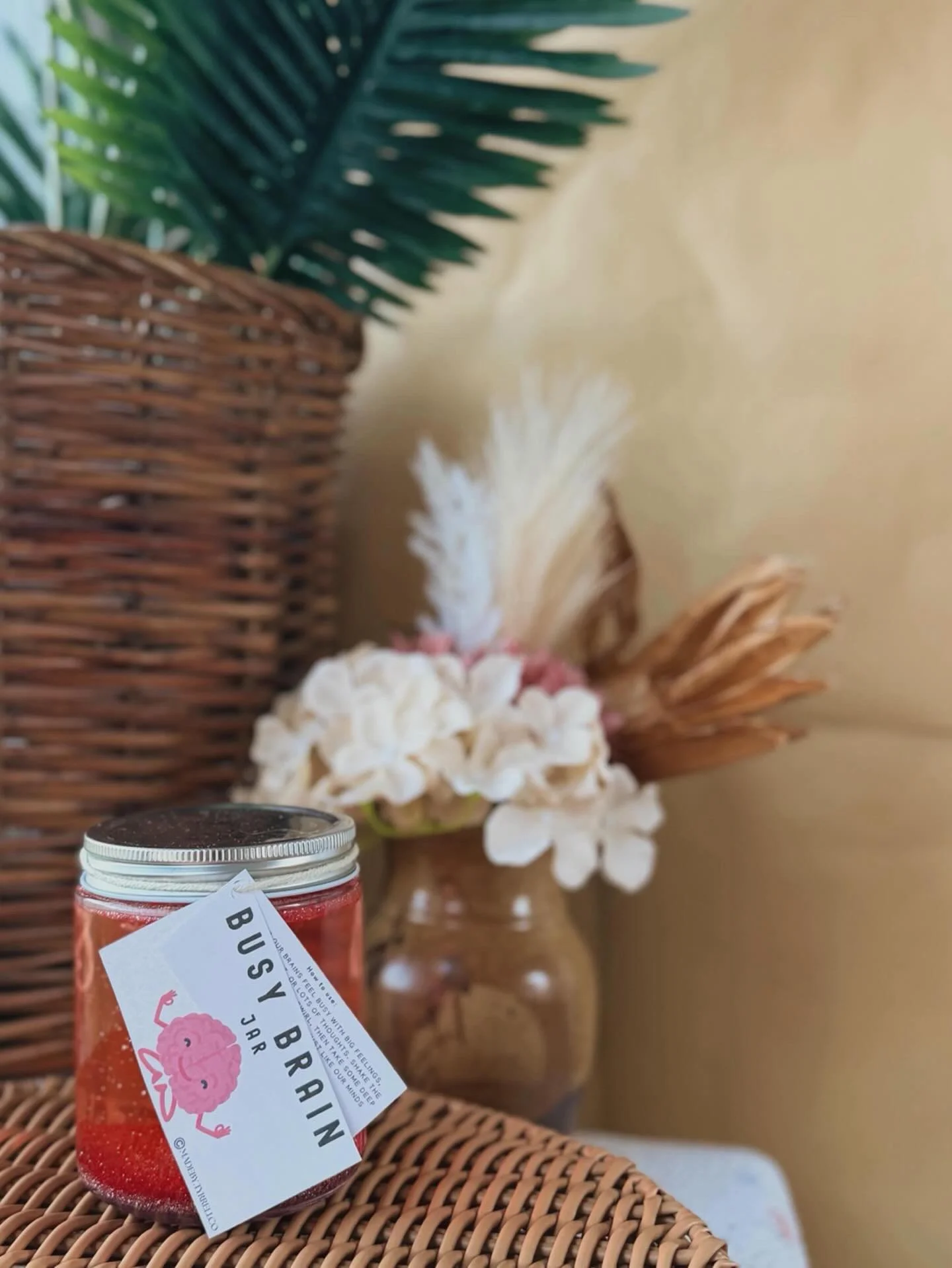 Close-up of a jar labeled 'Busy Brains JAR' with a pink brain illustration, set on a woven surface in front of a wicker basket, white flowers, and green palm leaves in a yellowish room.
