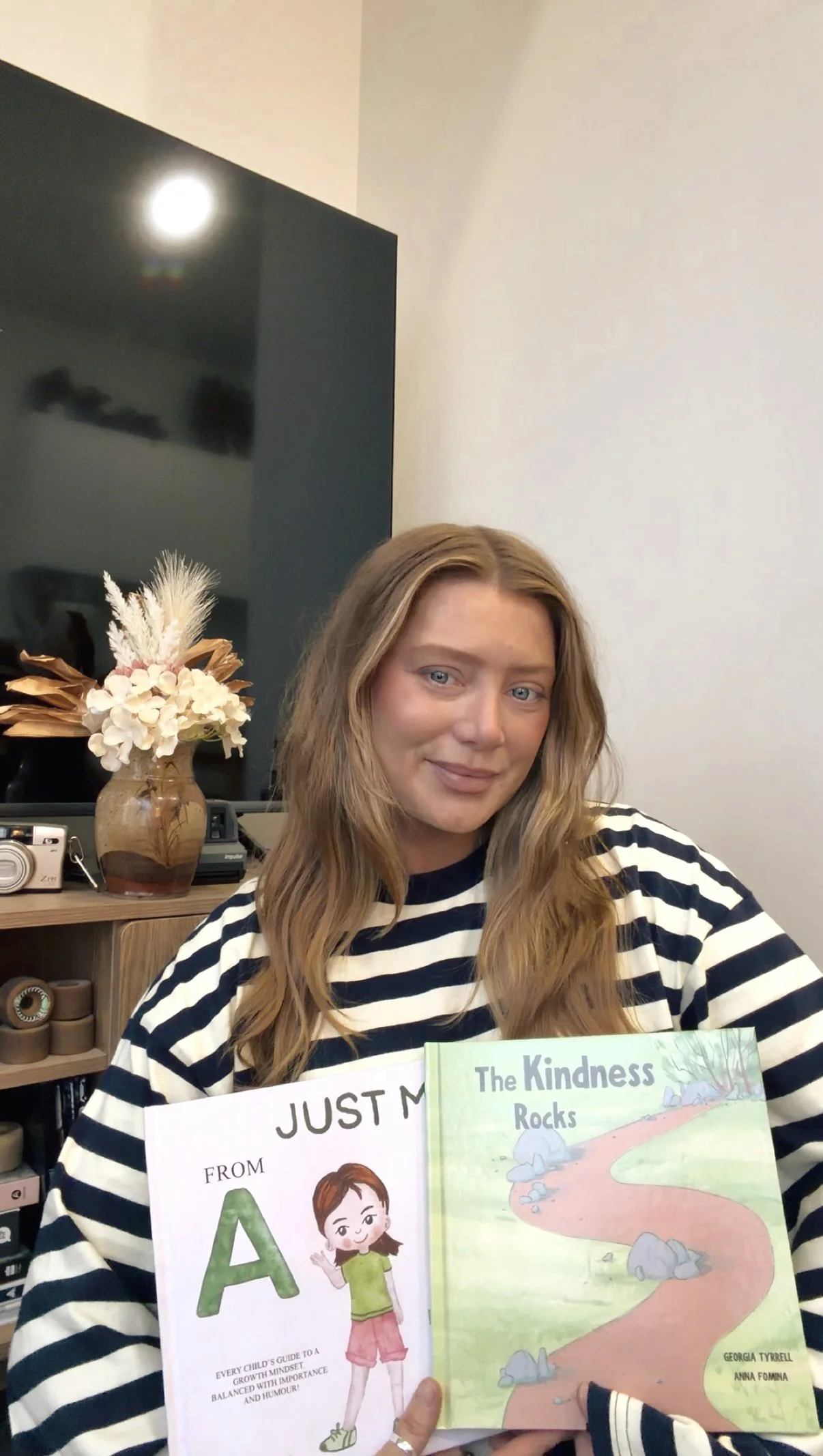 A young woman with long, wavy, light brown hair and blue eyes smiling while holding two children's books, one titled 'Just M' and the other 'The Kindness Rocks'. She is wearing a black and white striped shirt and is sitting in a room with a dark TV screen, a vase with white and blush pink flowers, a stack of rolls of tape, and a vintage camera on a wooden shelf in the background.