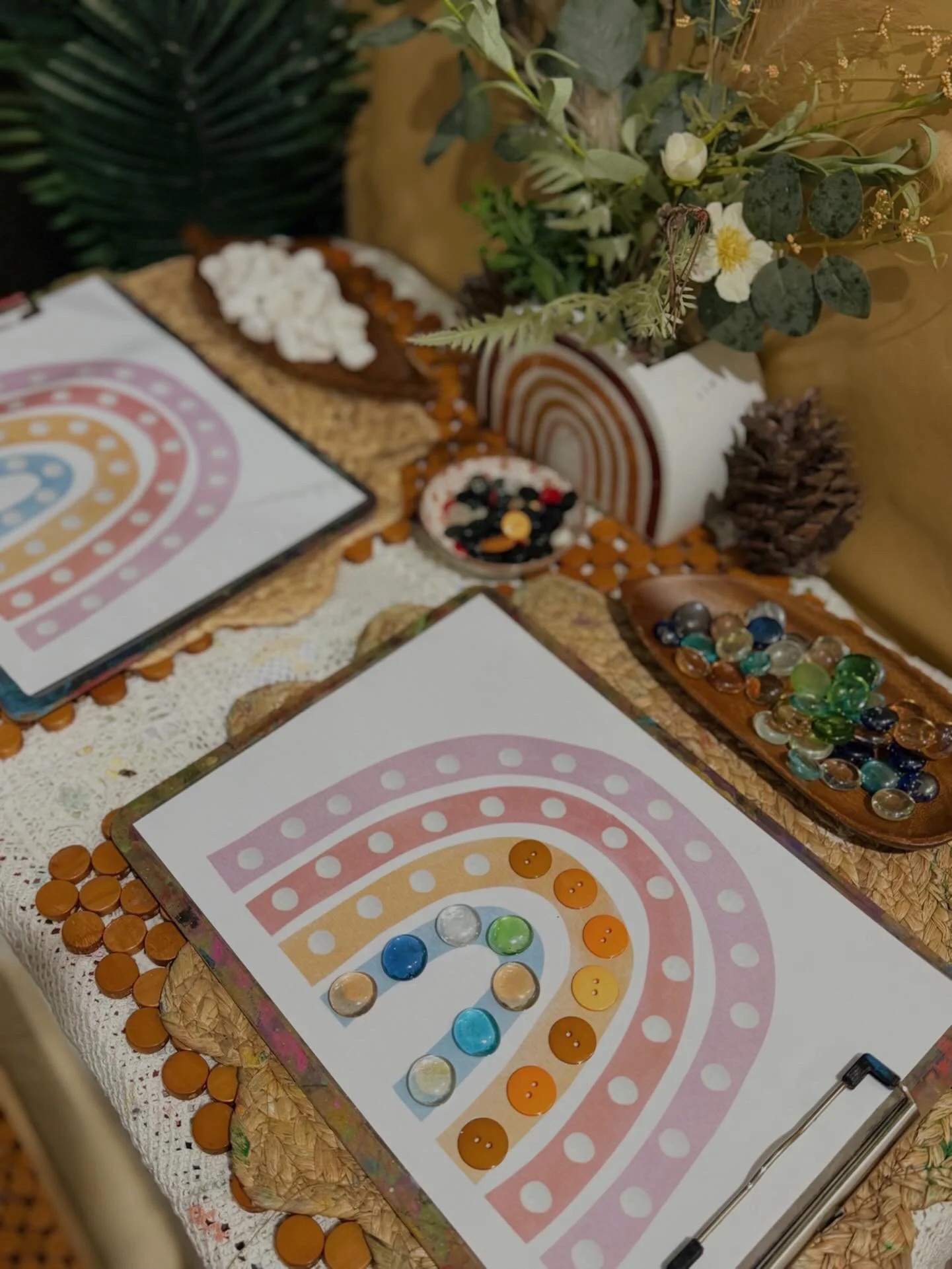 A board game with colored buttons arranged in a spiral pattern, placed on a table decorated with a white lace tablecloth and surrounded by decorative items including pine cones, paper flowers, and a plant with green leaves and white flowers.