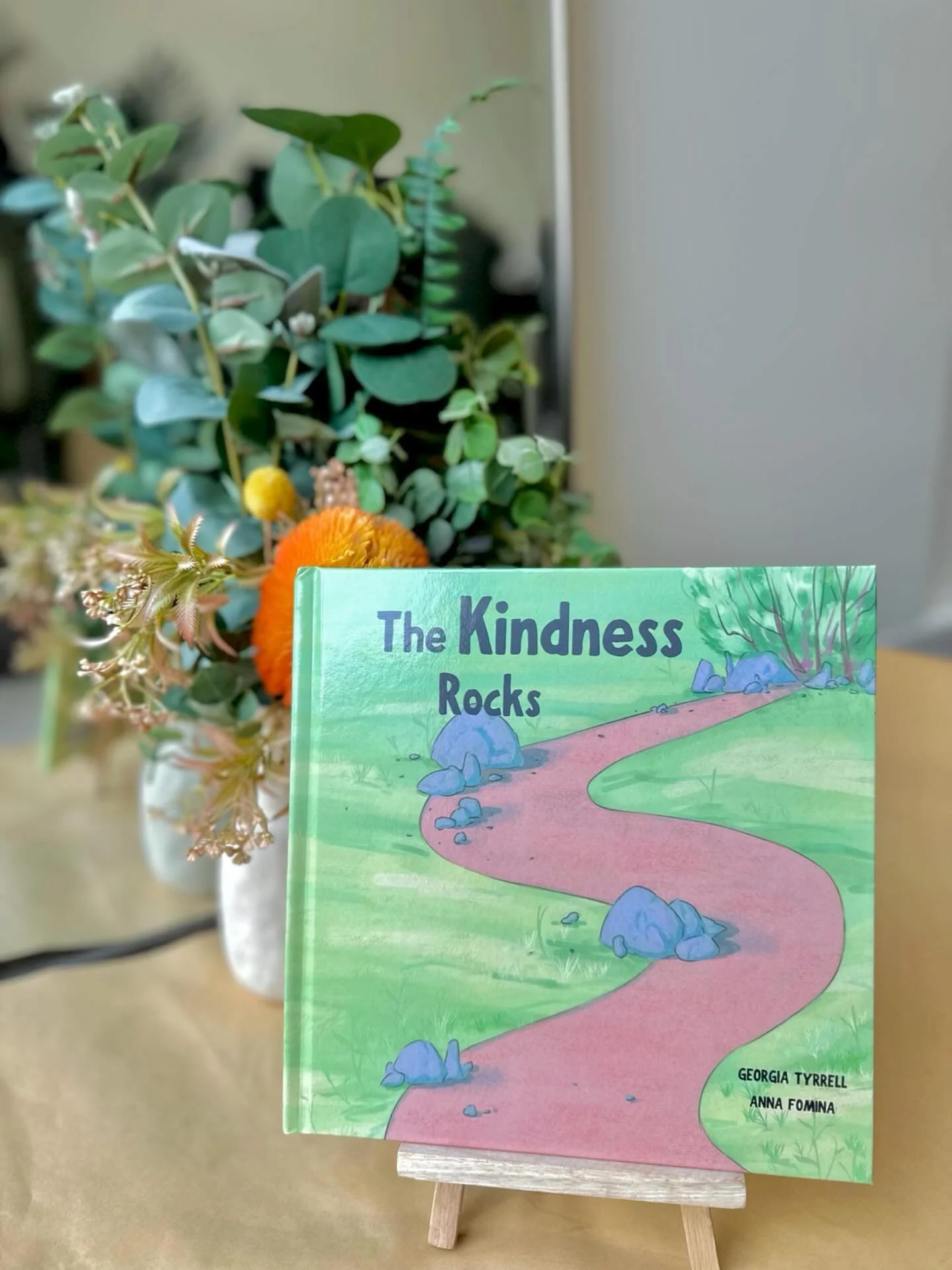 A children's book titled 'The Kindness Rocks' standing on a small wooden easel with a background of greenery and decorative flowers in a vase.
