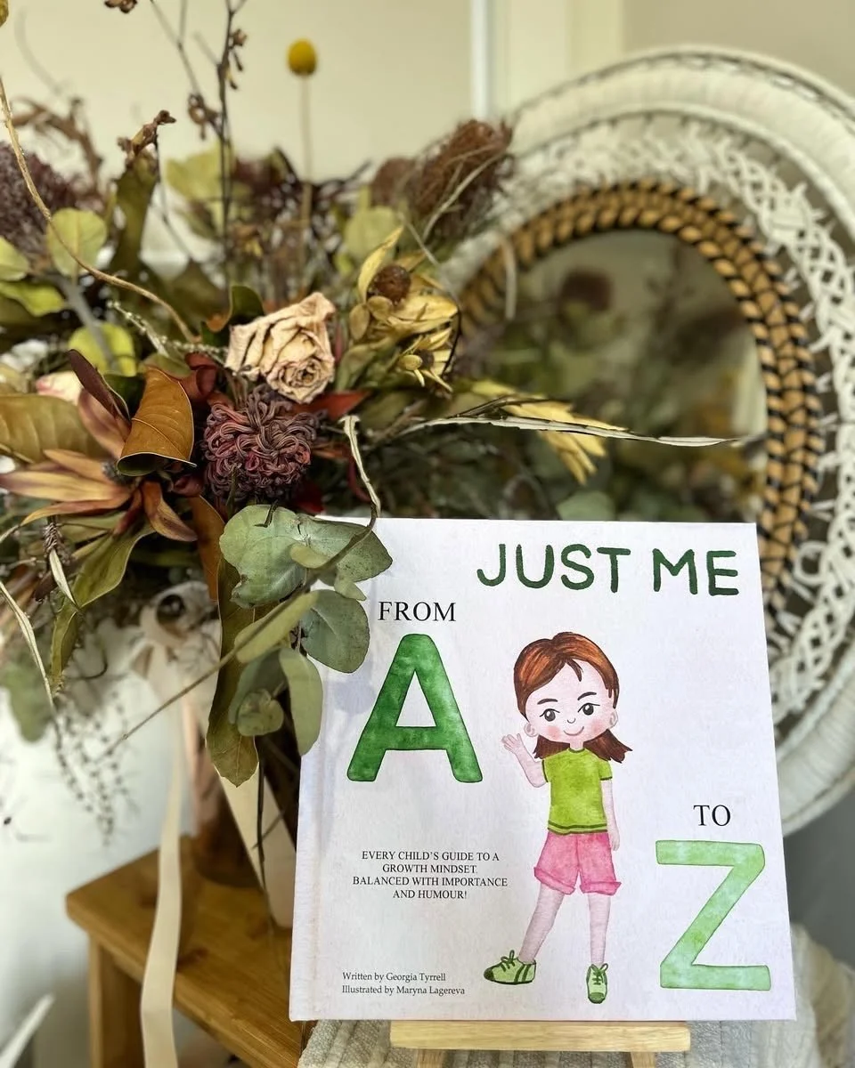 A children's book titled "Just Me from A to Z" featuring an illustration of a young girl in green shirt and pink shorts, waving, with dried flowers and leaves in the background