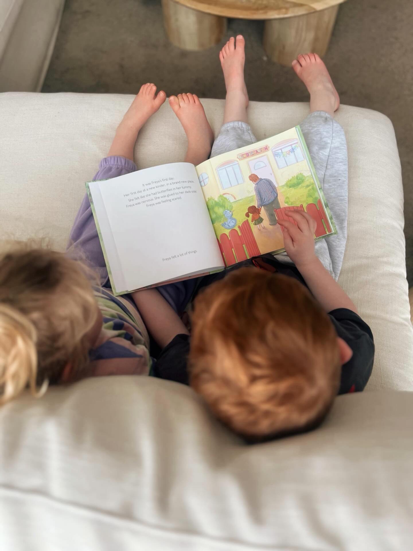 Two children sitting on a couch, one reading a children's book with colorful illustrations, while the other child sits next to them. The scene is viewed from above.
