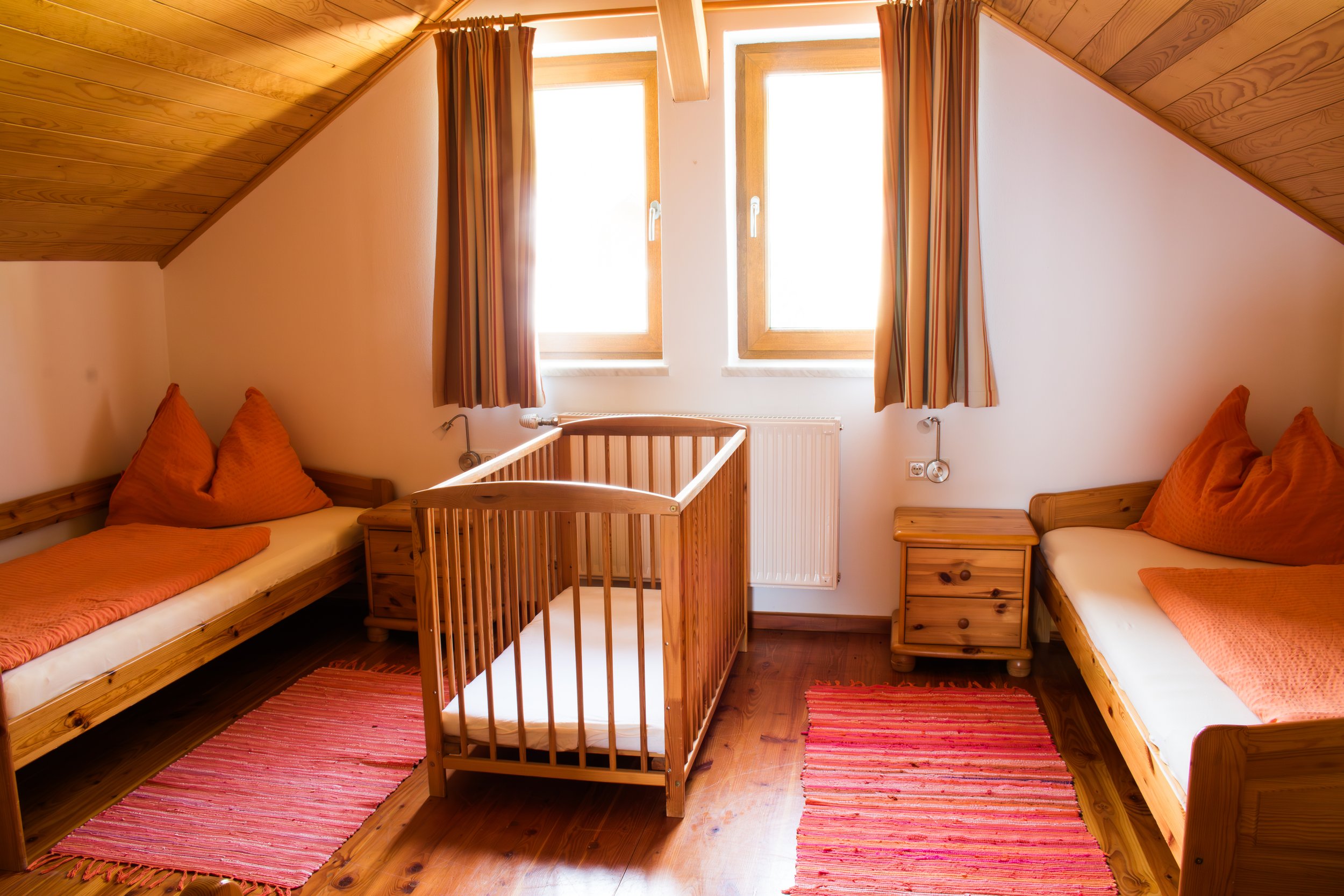 Holzverkleidetes Zimmer mit zwei Einzelbetten und einem Babybett, orangefarbene Kissen und Decken, rote Teppiche auf Holzboden, Dachschräge, zwei Fenster mit Vorhängen, Nachttischschränke aus Holz.