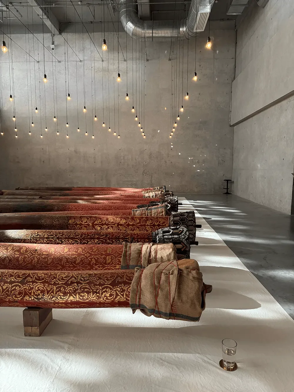 Large-scale installation on the second floor of Dib Bangkok featuring salvaged wooden logs arranged in rows beneath a grid of hanging bare light bulbs, set against raw concrete walls