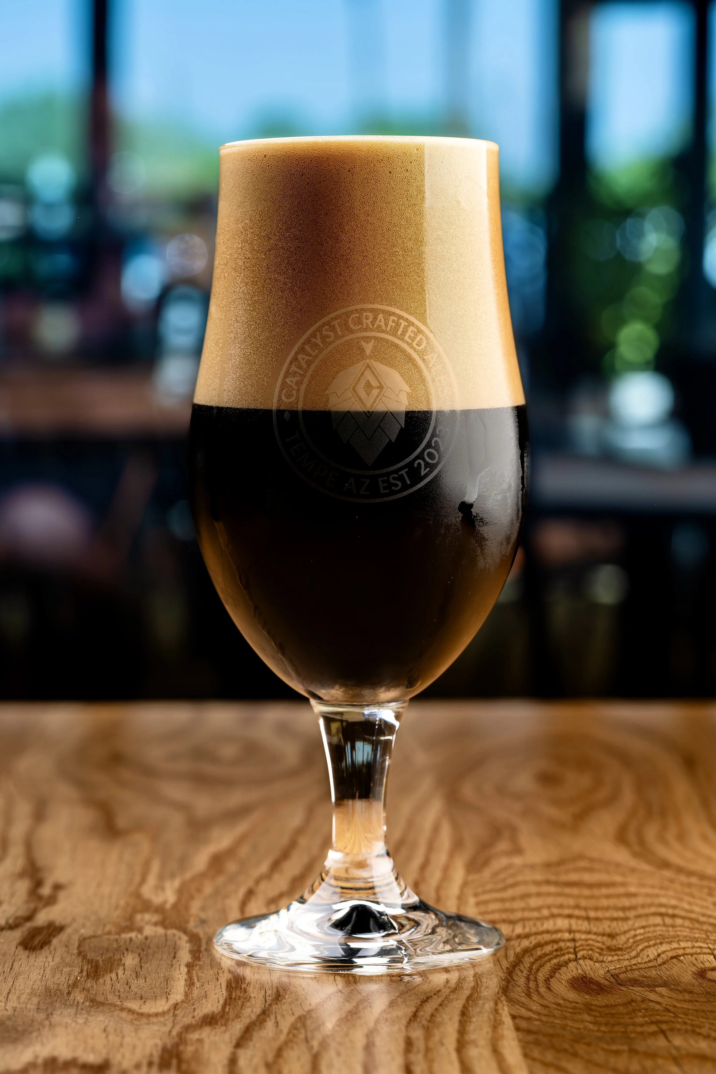 A close-up of a glass filled with a dark craft beer, with a head of foam and a logo etched on the side.