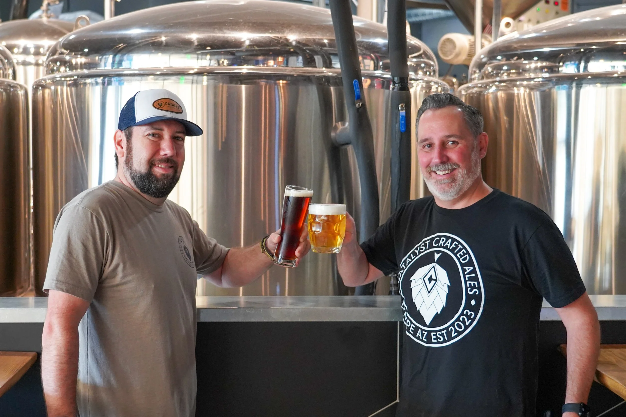 Two men standing in front of shiny stainless steel brewing tanks inside a brewery, raising their glasses in a celebratory toast.