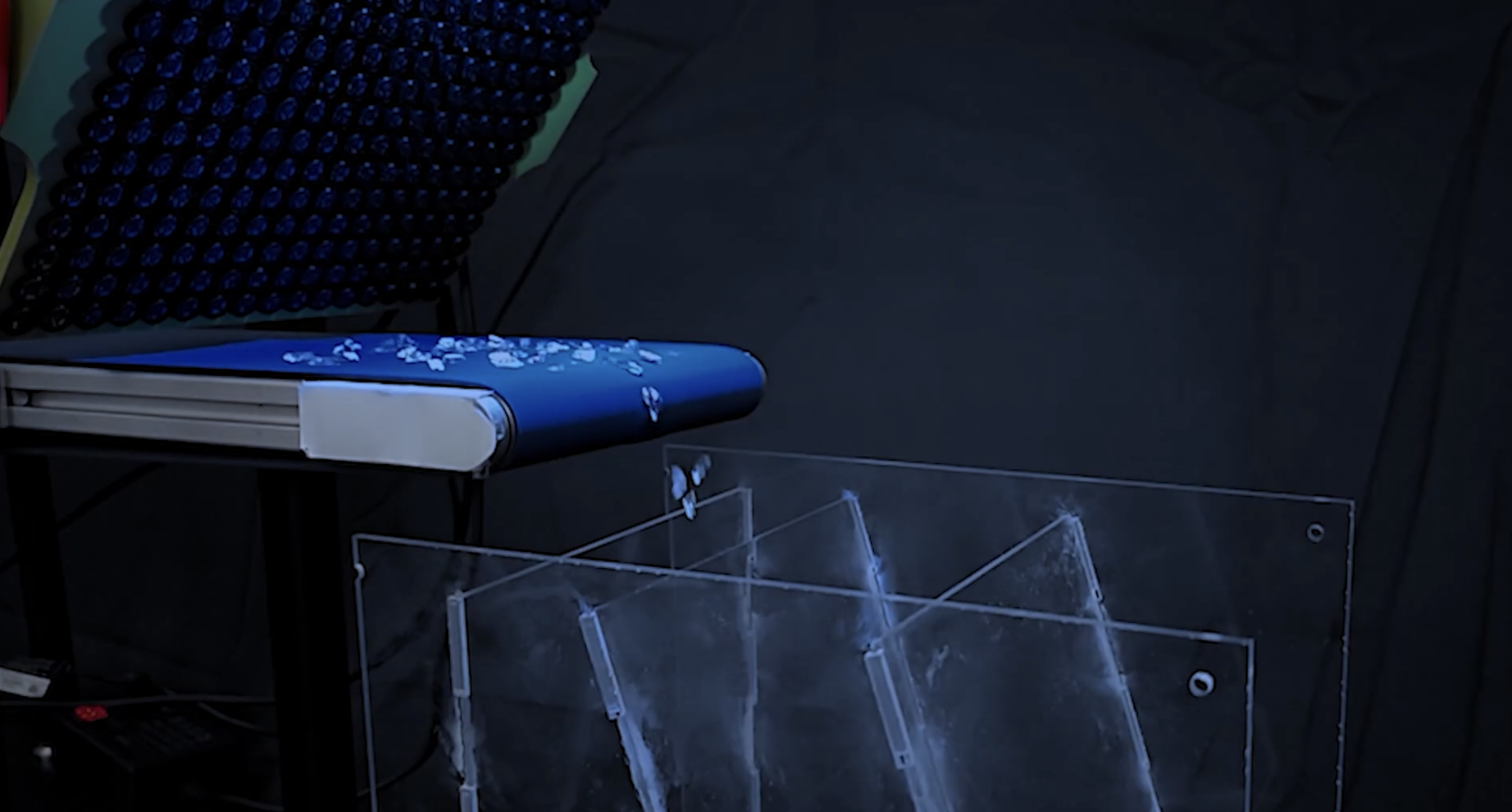 A display of a digital scientific instrument with small white particles on top, placed on a blue surface. Several transparent acrylic stands with illuminated edges are positioned nearby, and the background is dark.