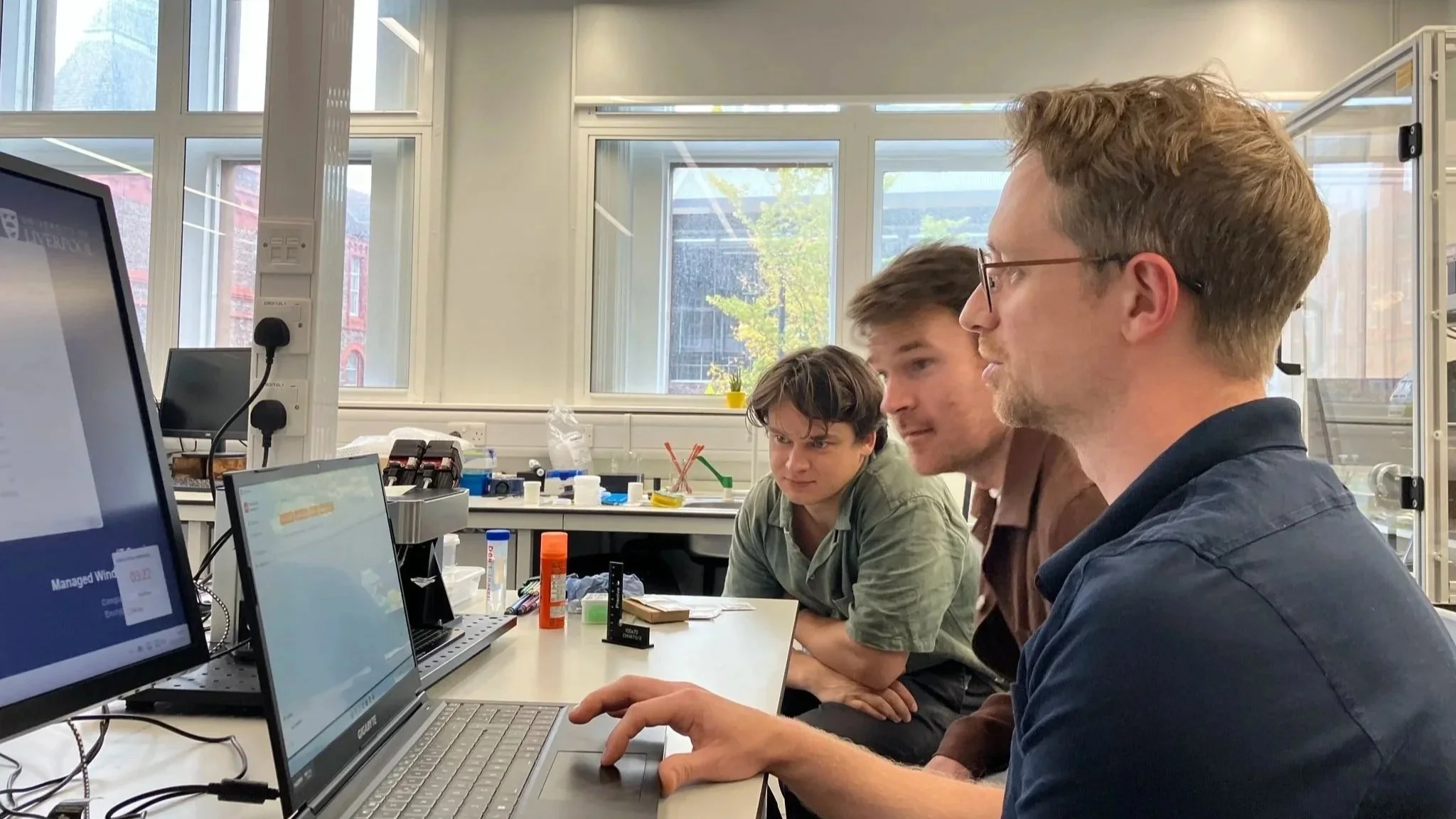 Three young men working together at a science laboratory desk with computers, laboratory equipment, and test tubes, in a bright room with large windows.