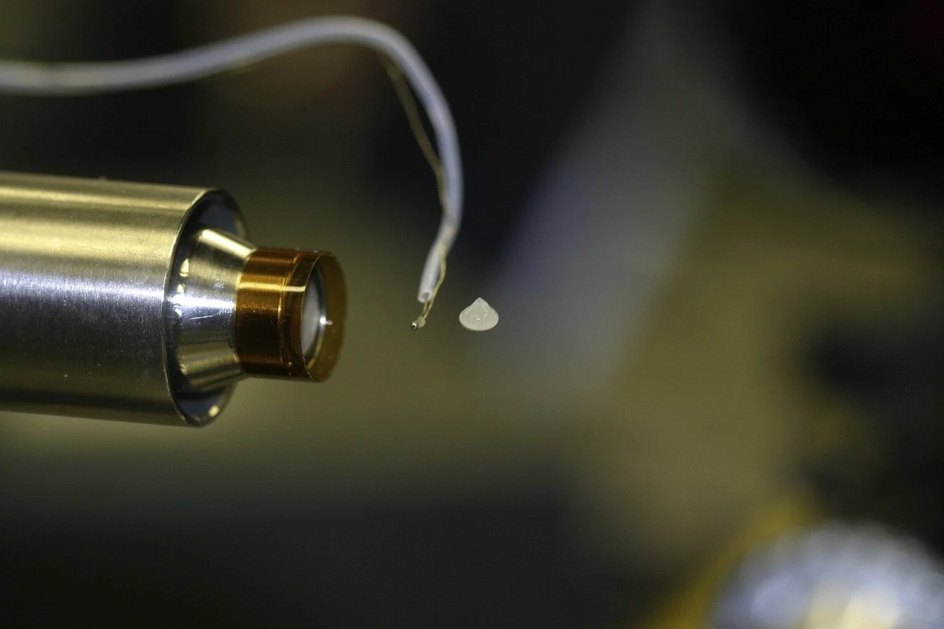 Close-up of a scientific instrument with a metallic cylindrical component and thin wires, with a small, shiny, irregularly shaped white object suspended nearby.