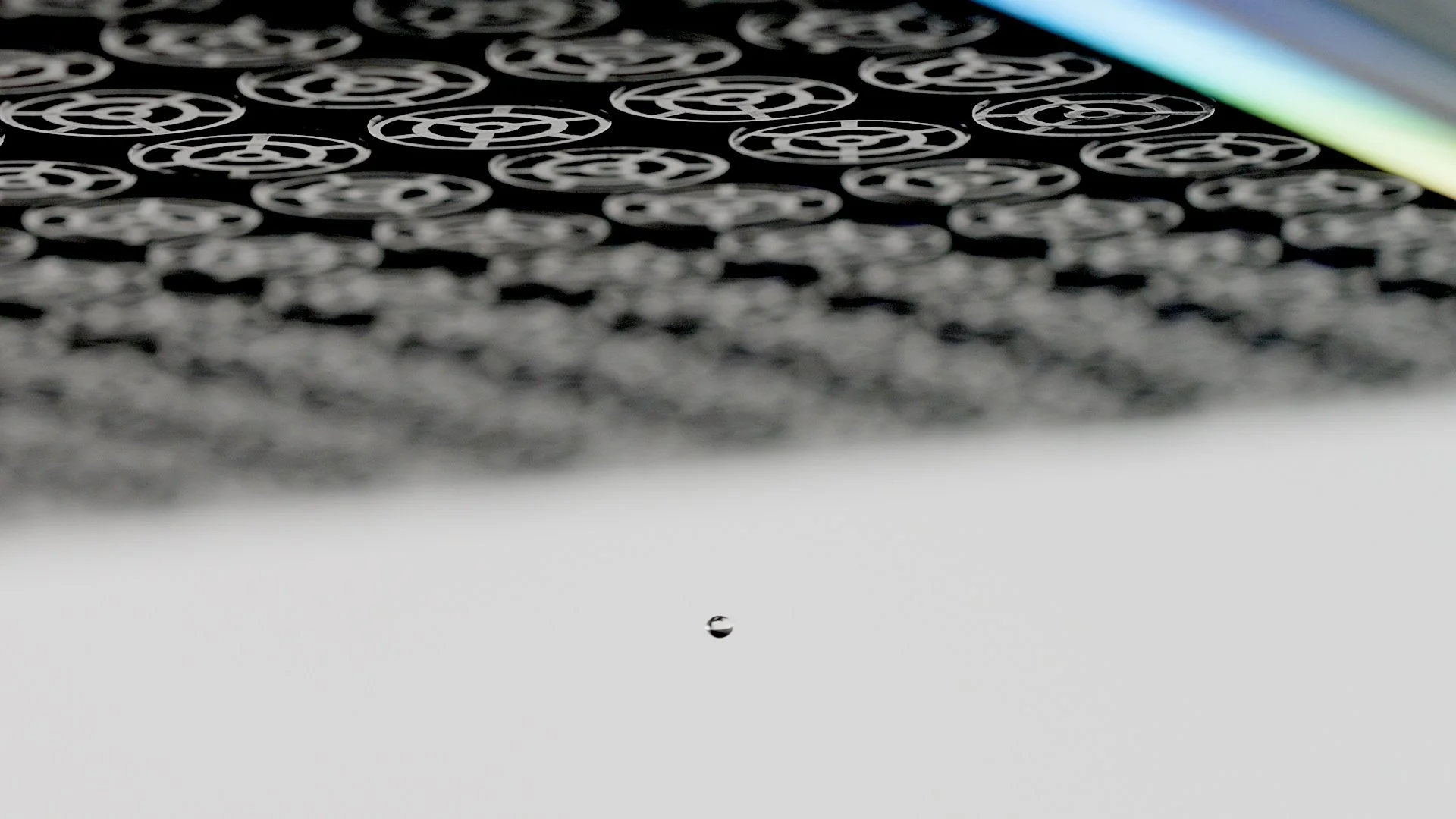 Close-up of small droplets of water on a surface with a pattern of circular designs, with colorful striped light reflections in the background.