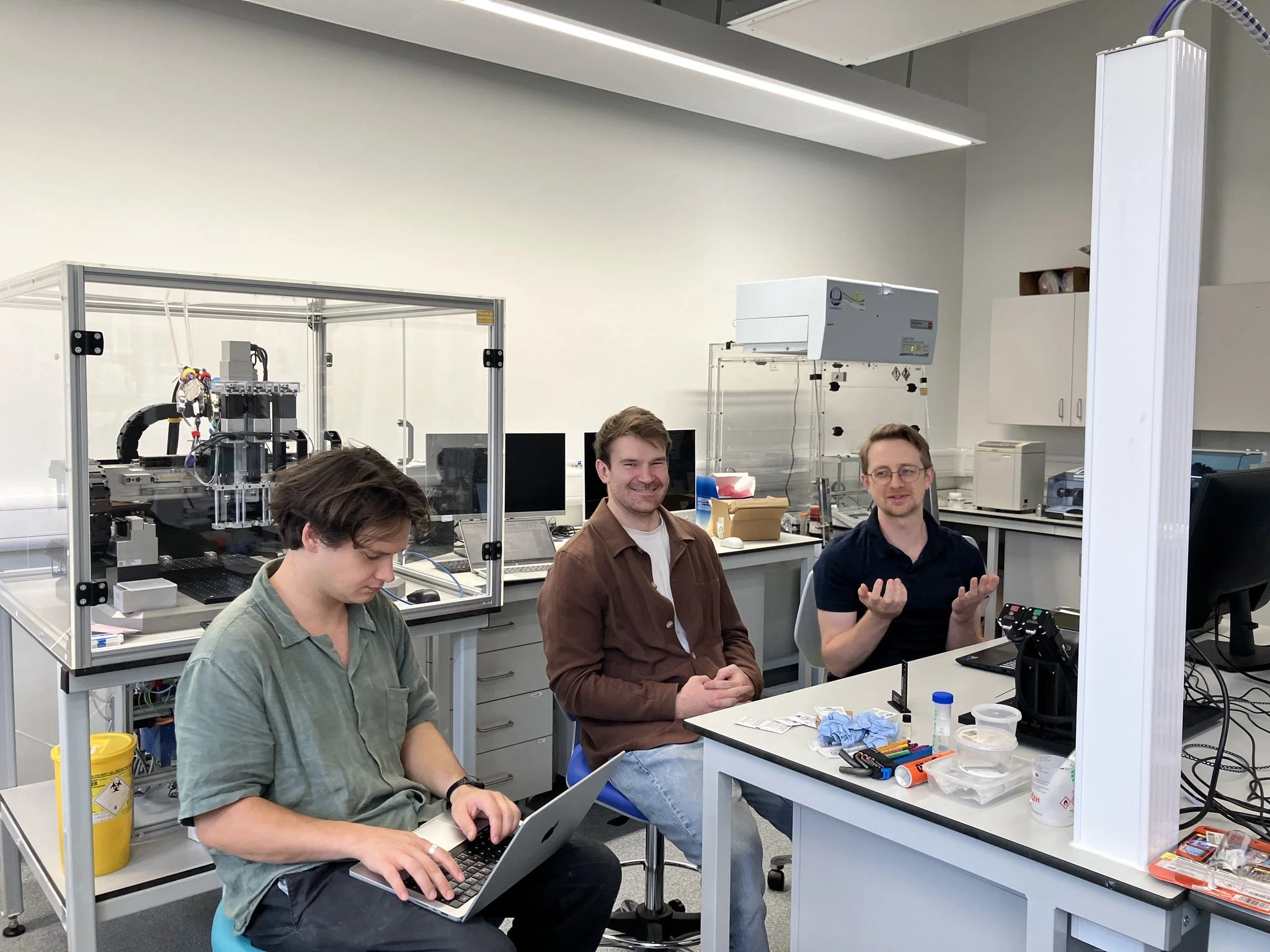 Four people working in a laboratory with scientific equipment and computers. One person is typing on a laptop, and two men are seated, engaged in conversation. The workspace is filled with lab tools, wires, and supplies.