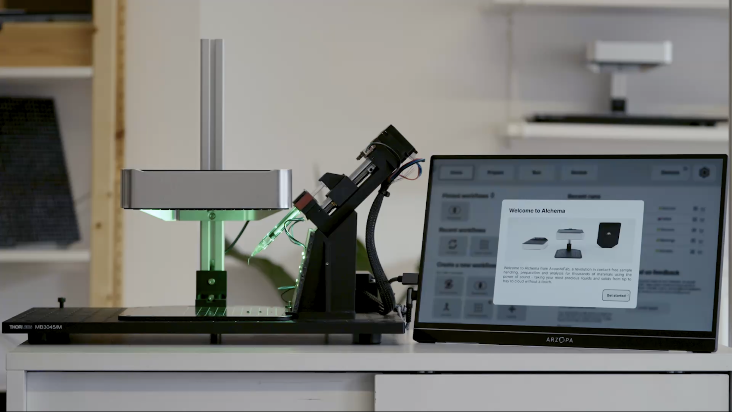 Laboratory setup with a computer screen displaying a welcome message for 'Alchema' and a device with a syringe and electronic components, possibly for biochemistry or scientific research.
