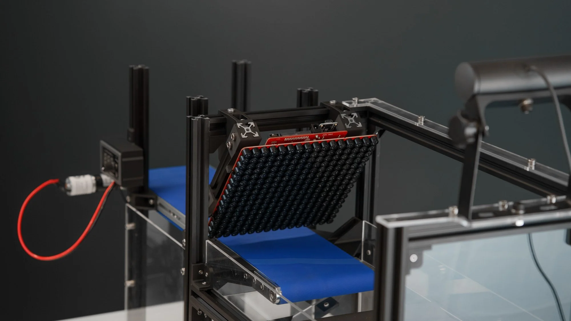 A mechanical setup with a black frame holding a grid of small black batteries, connected to wires, on a blue conveyor belt against a plain dark background.