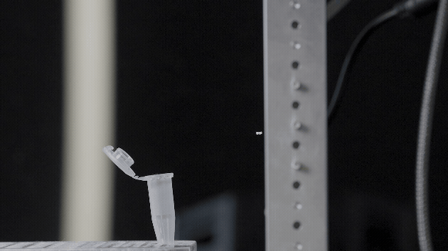 Close-up of a small plastic or glass object with a tap or spout, possibly a laboratory or industrial equipment, mounted on a table or surface with a dark background.