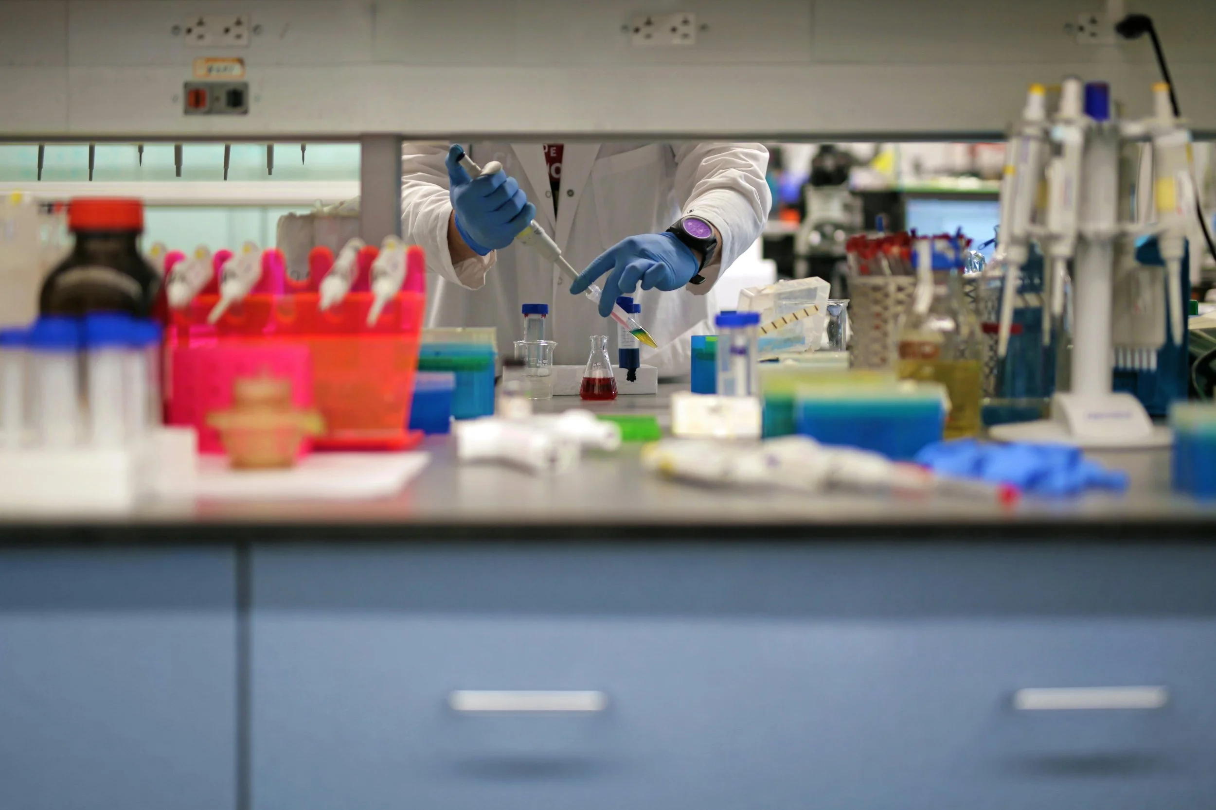 A scientist in a laboratory is using a micropipette to transfer a liquid into a small container, surrounded by various laboratory equipment and supplies.