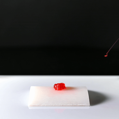Close-up of a small, red gummy candy on a white surface, with a black background and a red string or antenna visible in the upper right corner.
