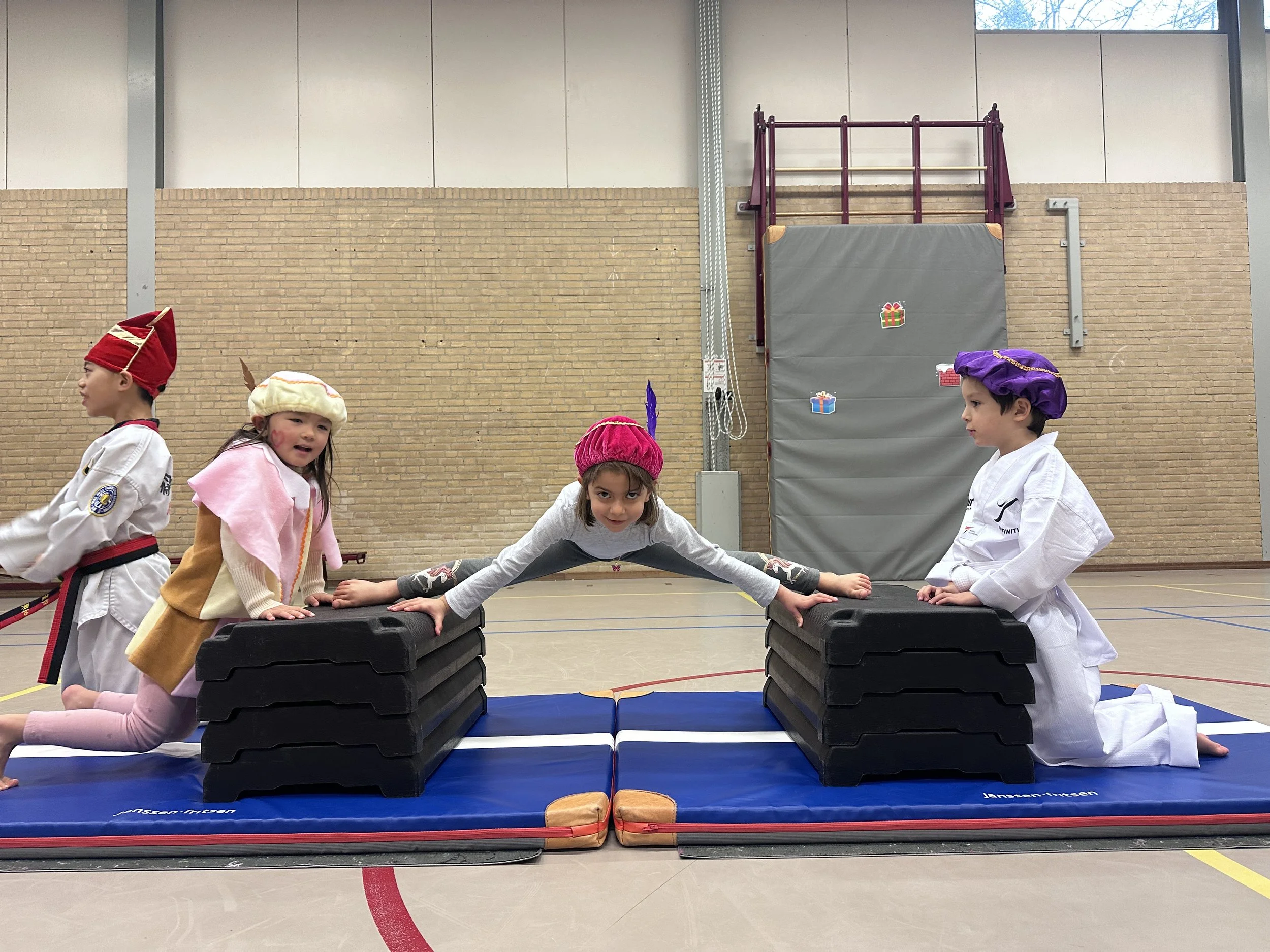 Three children in martial arts uniforms balancing between chairs; one in the center doing a split and the others on the sides.