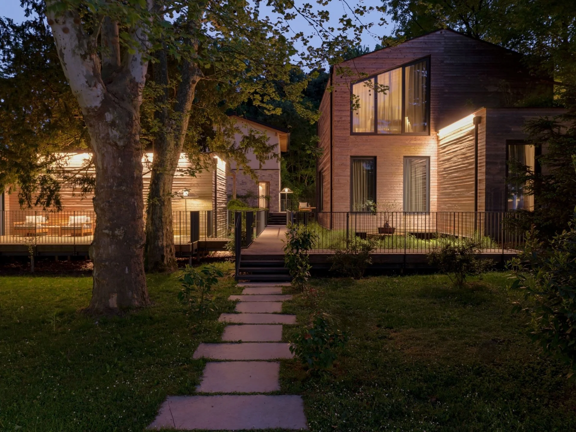 Casa moderna in legno circondata da alberi, illuminata durante il crepuscolo, con un sentiero di pietre nel prato frontale.