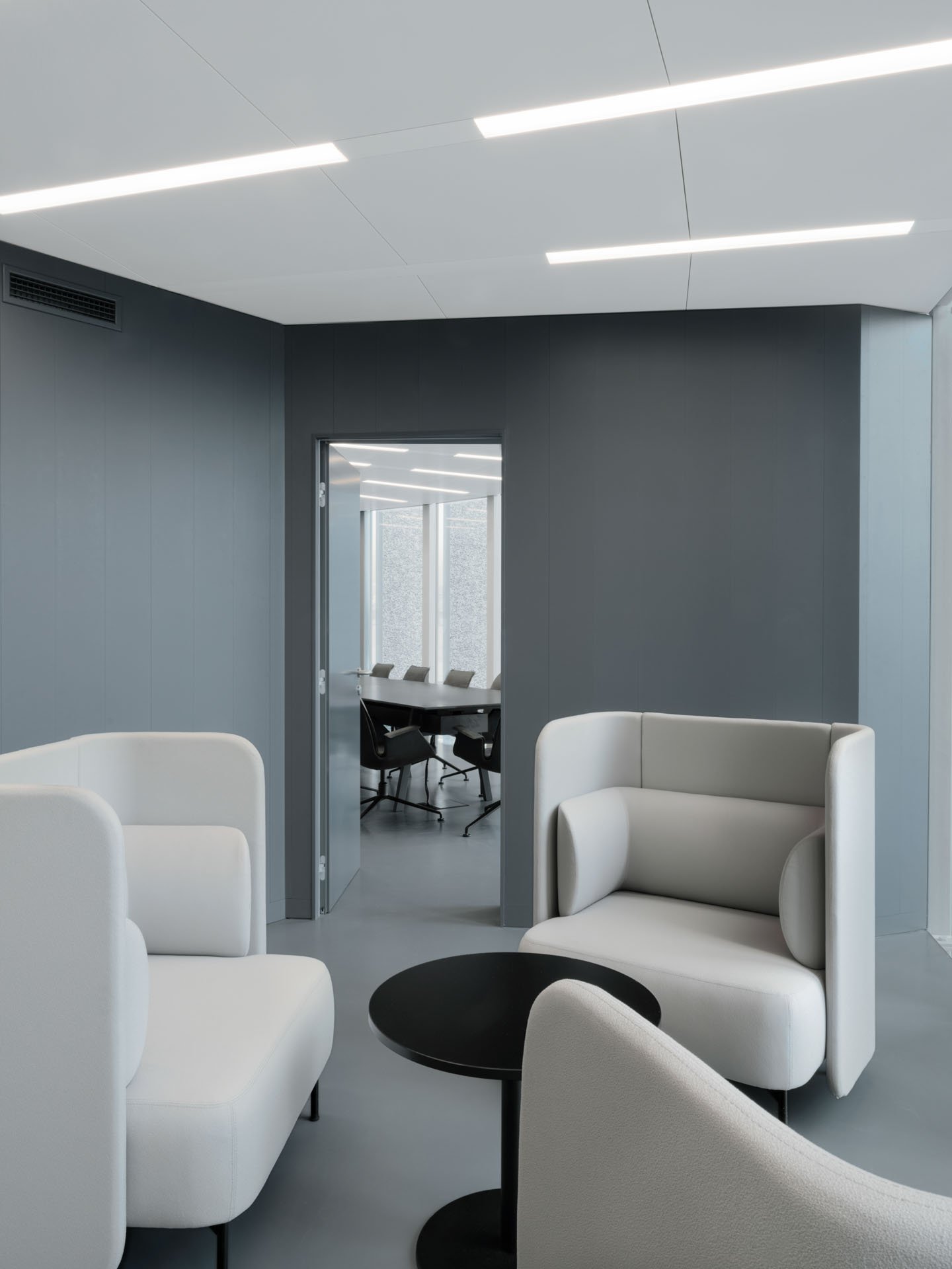 Interno di ufficio moderno con sedie bianche, tavolini neri e pareti grigie. Si vede una stanza con una grande finestra e un tavolo con sedie in uno sfondo.